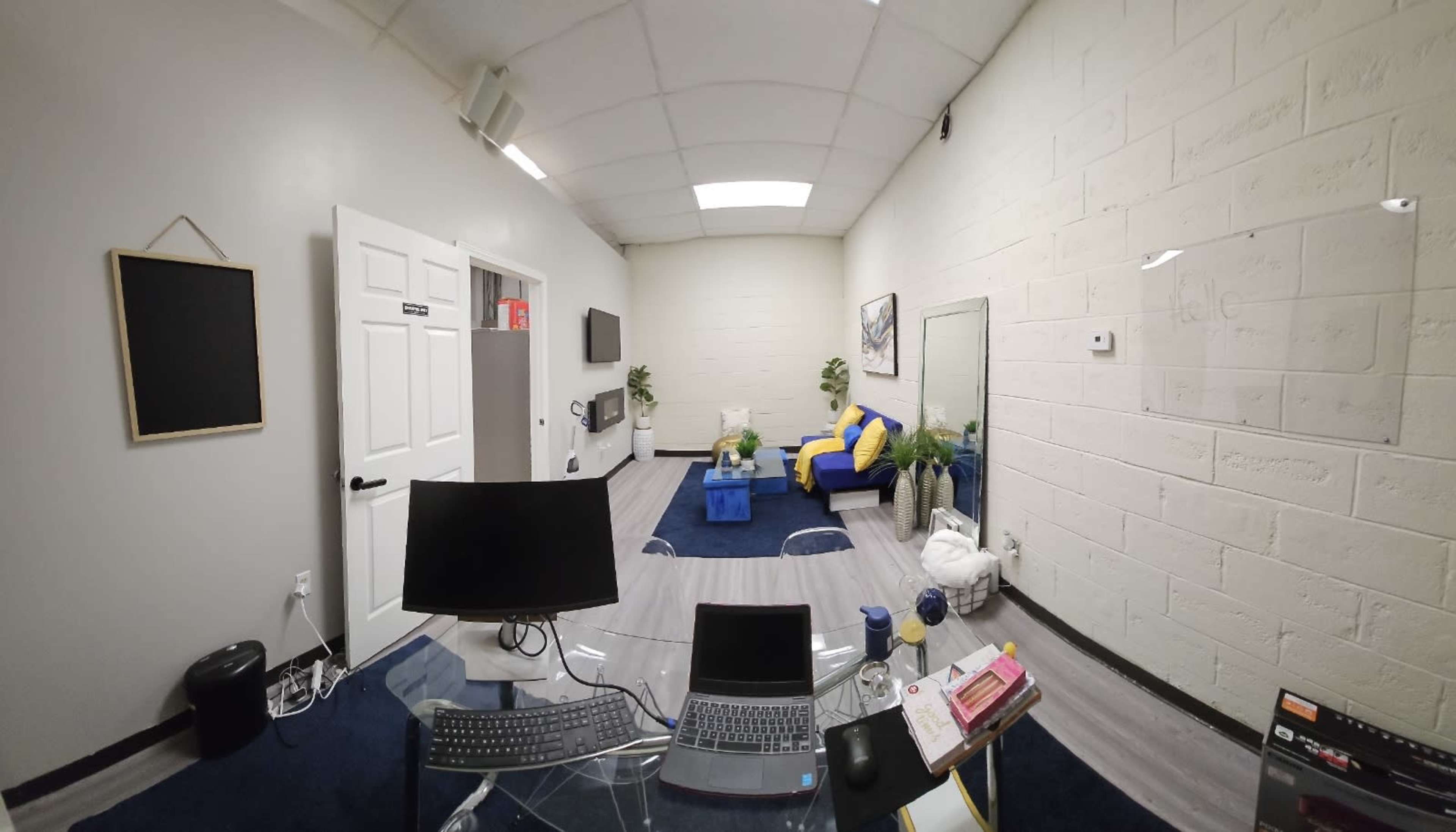 The image shows a small office space featuring a desk with a laptop, a couch, a blue table, and decorative plants on the floor.