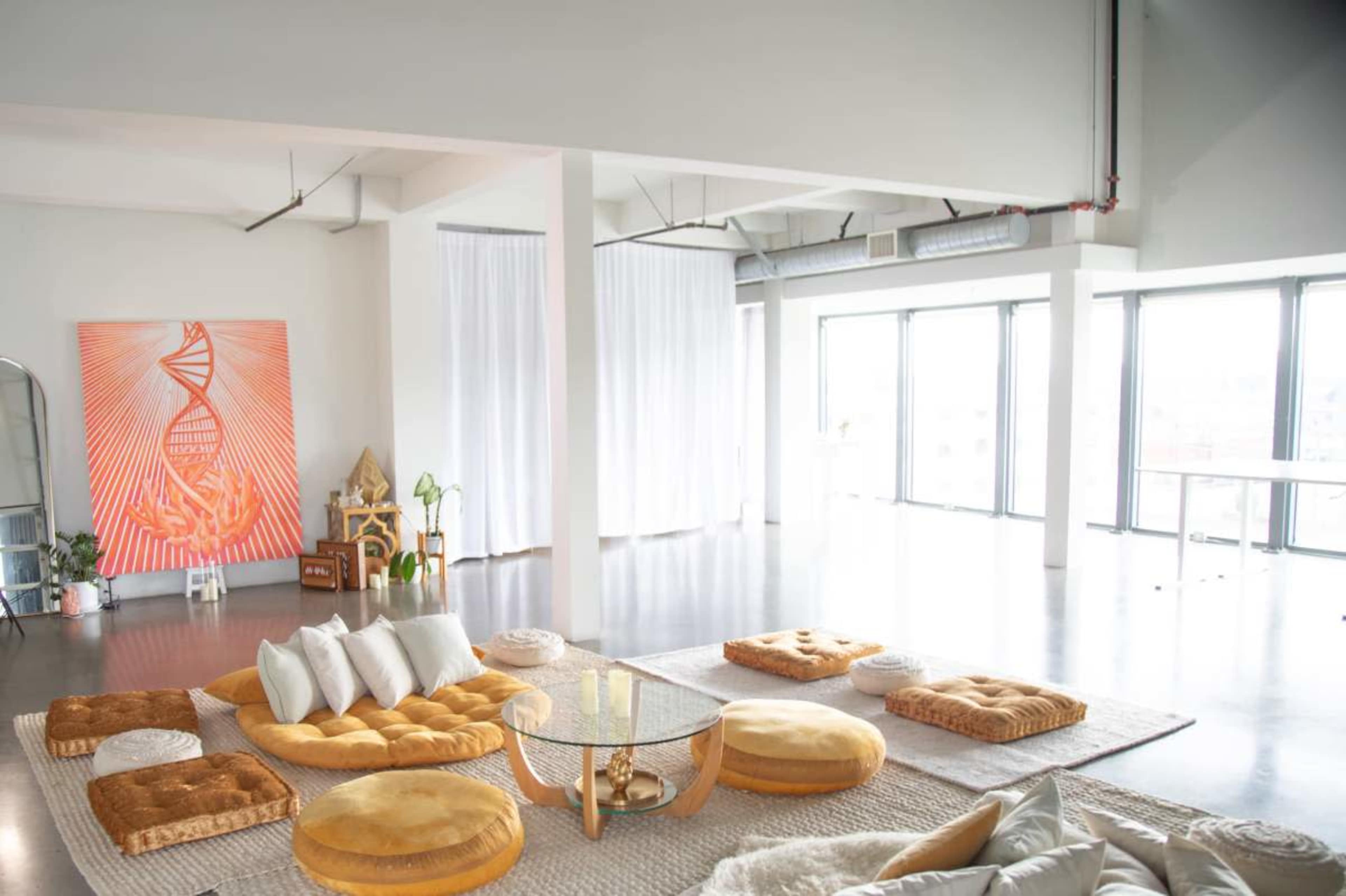A spacious, modern room filled with various cushions and seating arrangements, large windows letting in natural light, and a vibrant artwork on the wall.