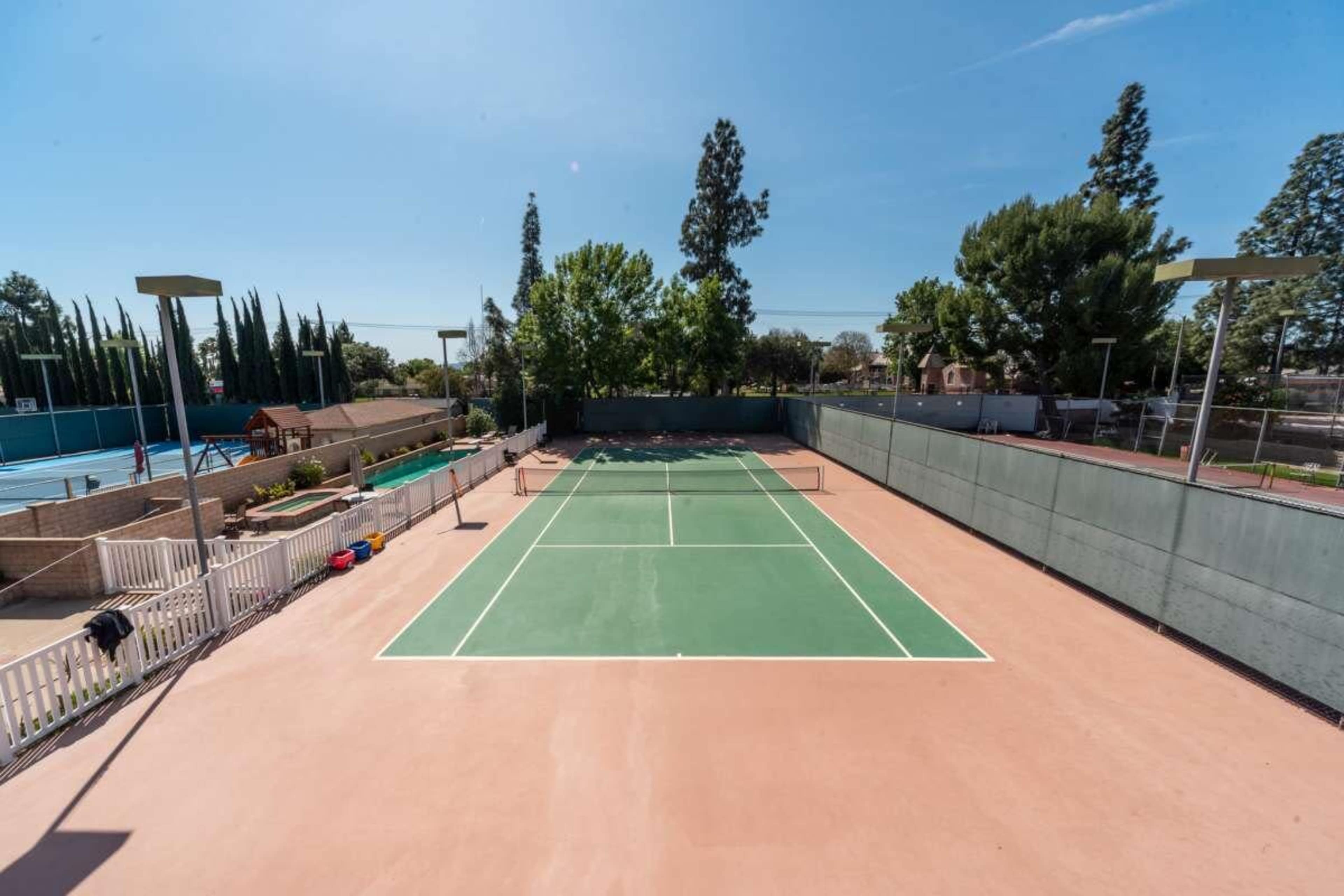 The image shows two tennis courts, one clay and one hard surface, surrounded by trees and a clear blue sky.