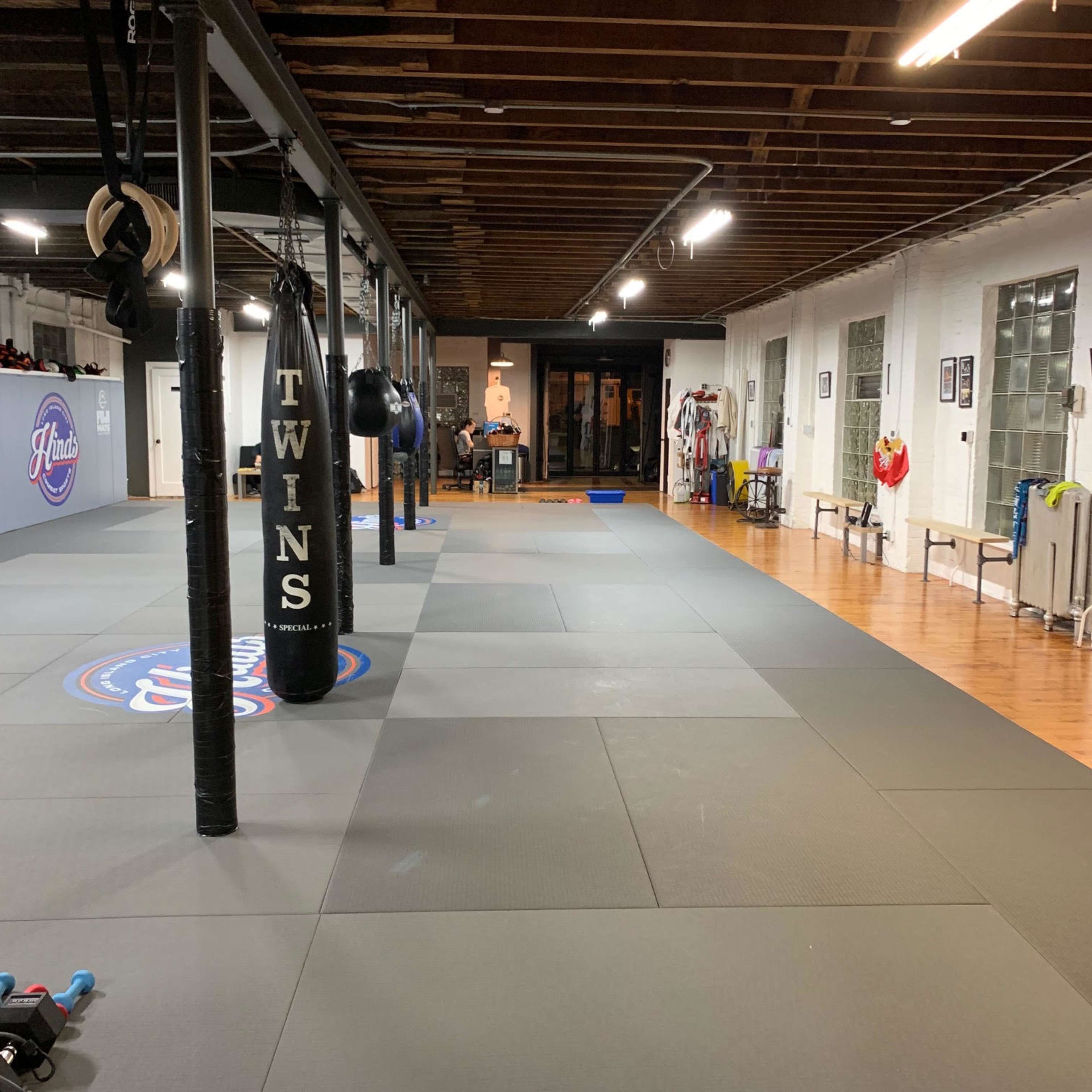 An empty martial arts gym with gray mats, black punching bags hanging from the ceiling, and various training equipment along the walls.