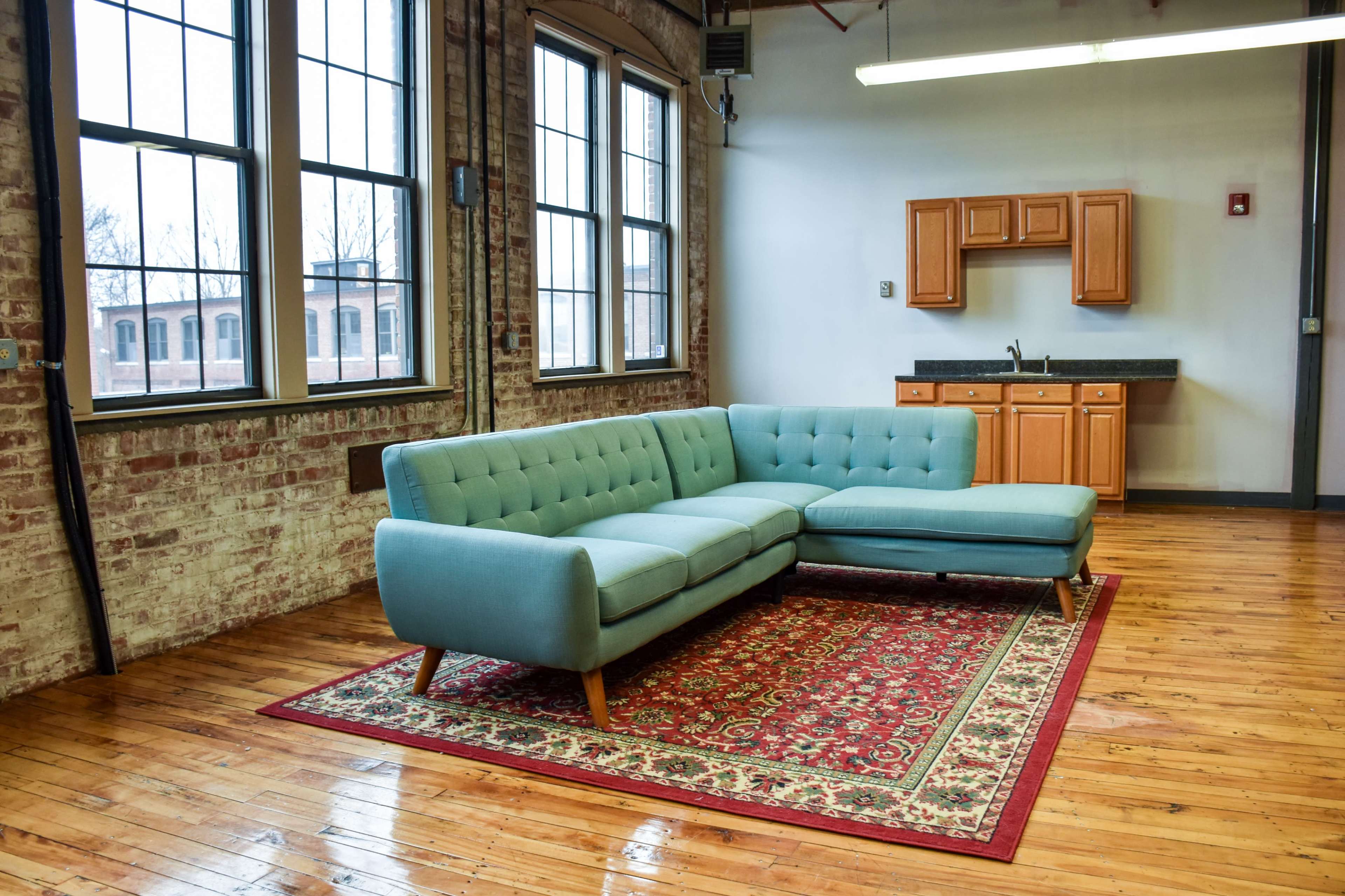 A light blue sectional sofa sits on a patterned rug in a room with exposed brick walls and large windows, adjacent to a kitchen area with wooden cabinets.