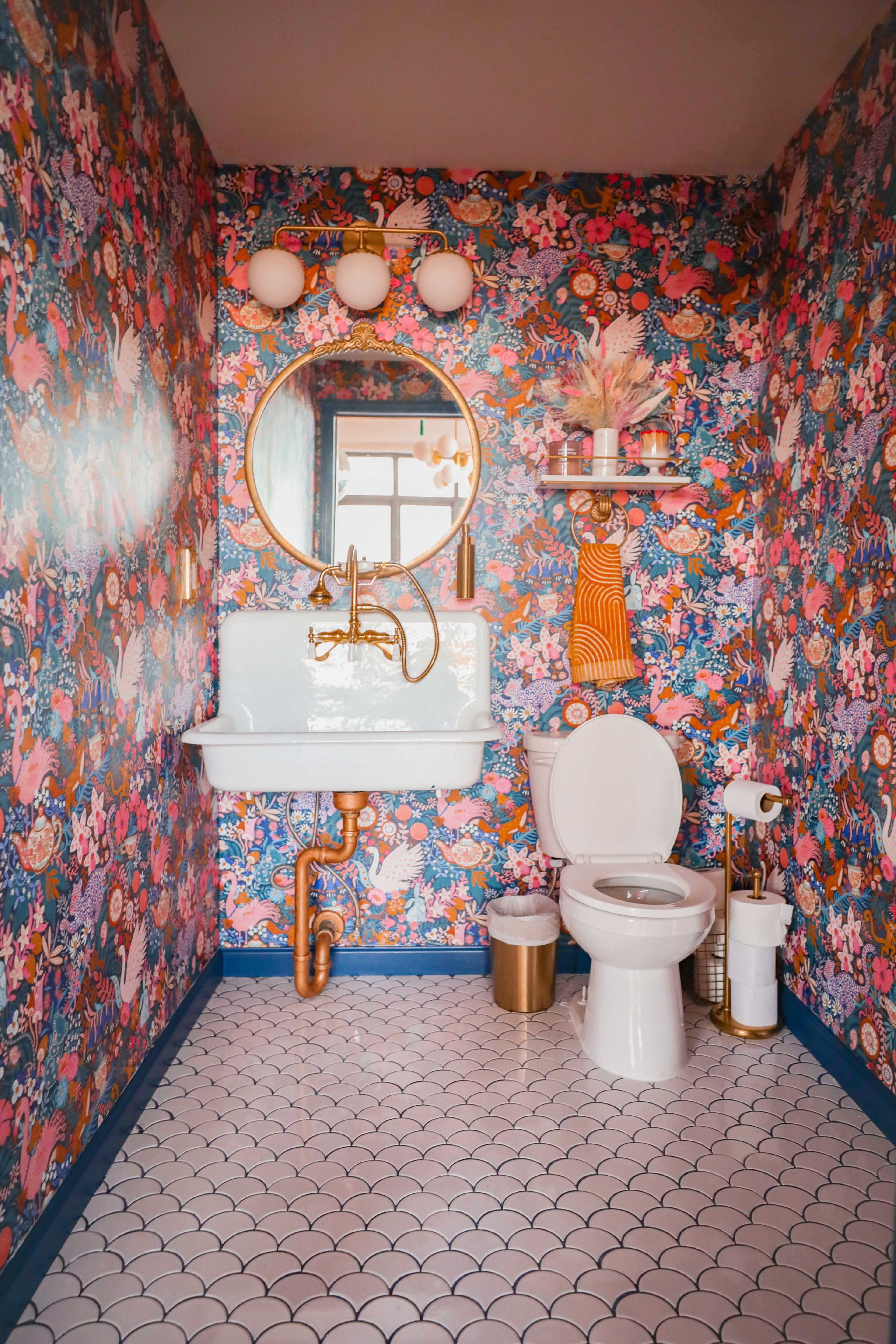 A colorful bathroom features floral wallpaper, a round mirror above a blue sink, and a gold faucet, along with a white toilet and patterned floor tiles.
