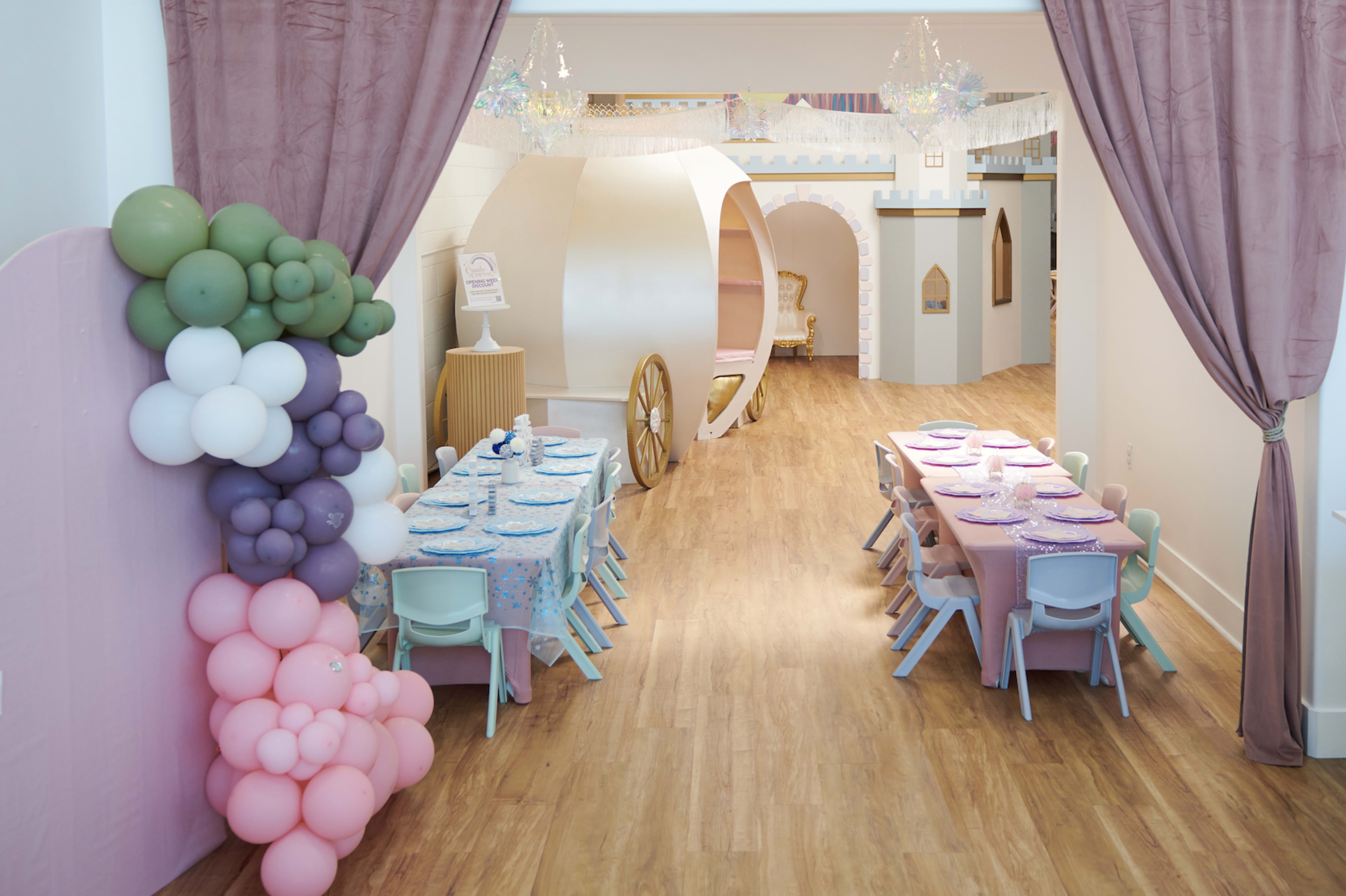 The image shows a colorful children's party area with two tables set for a celebration, surrounded by decorative balloons and whimsical architectural elements.
