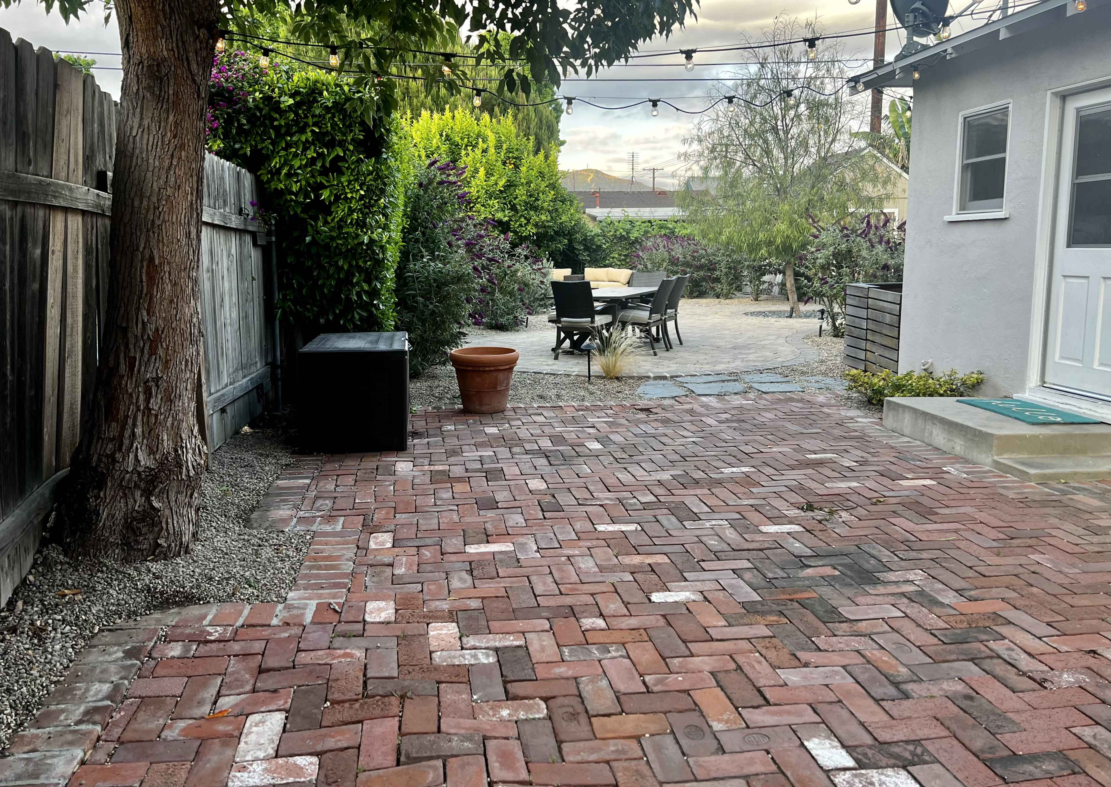 A brick patio with a black storage box and a potted plant is surrounded by greenery and outdoor furniture in a backyard.