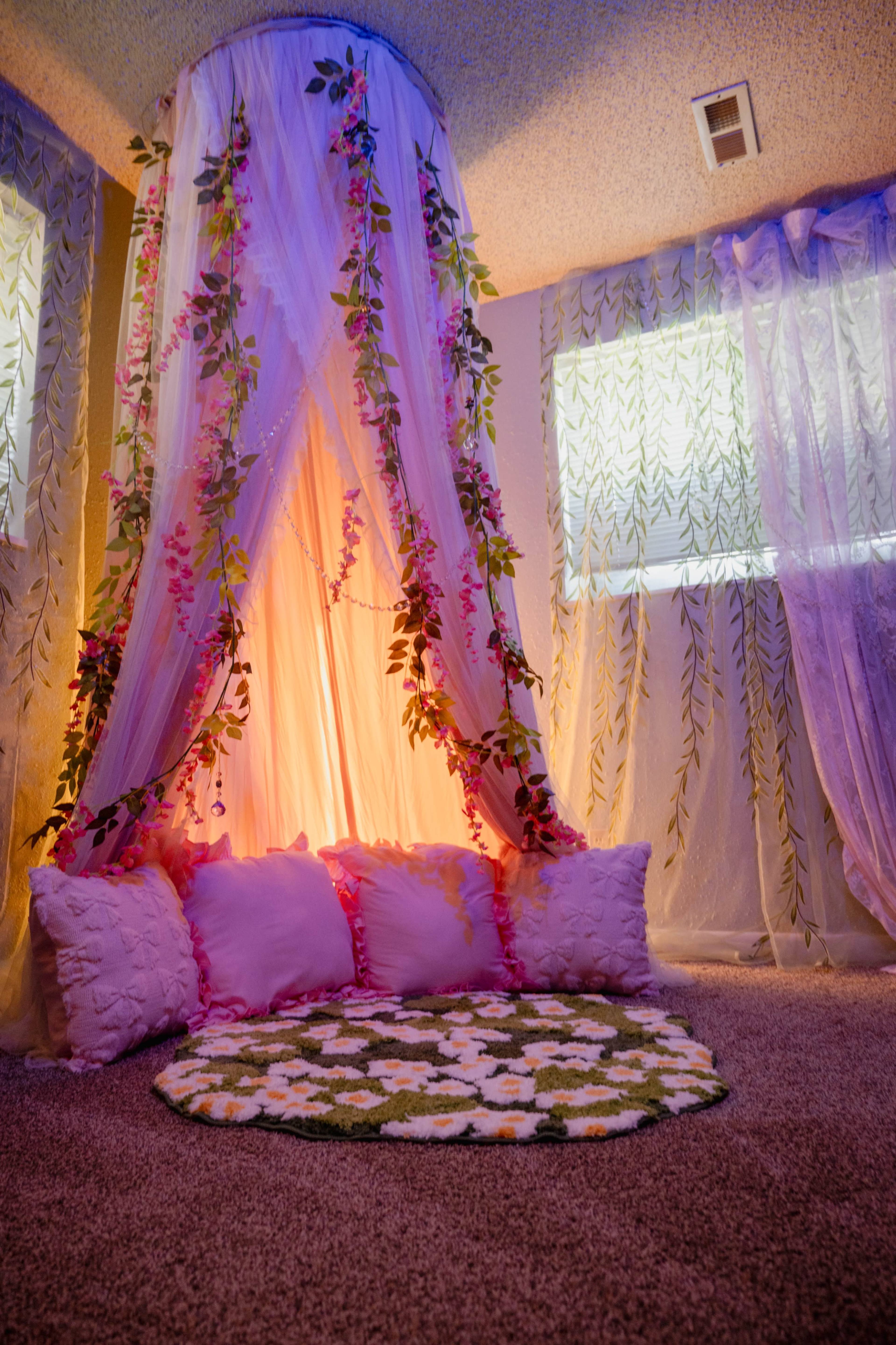 The image shows a cozy corner with a fabric canopy adorned with floral decorations, surrounded by cushions and a circular floral rug on a carpeted floor.