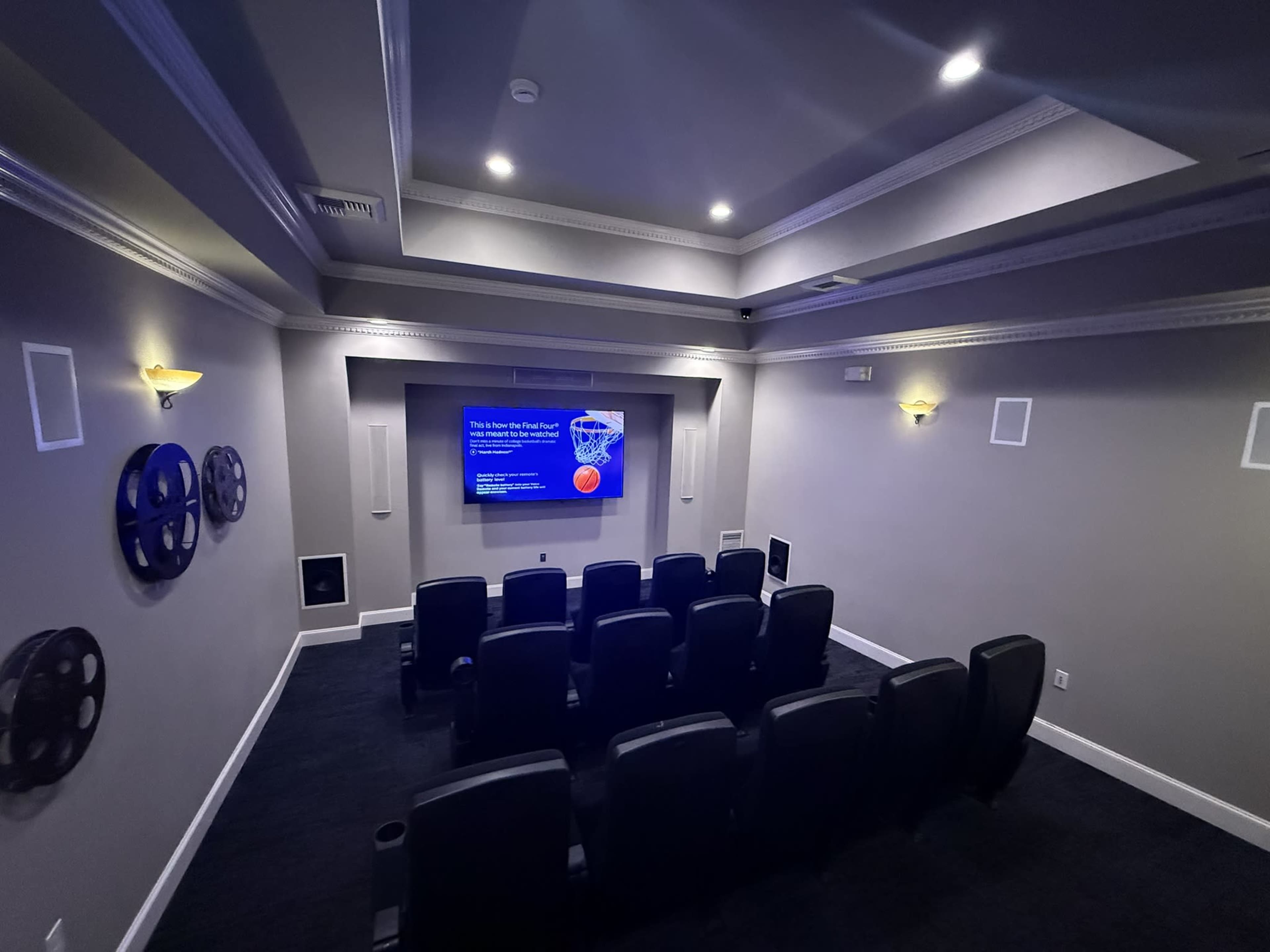 The image shows a home theater with tiered seating, a large screen displaying a presentation, and decorative film reel wall art.