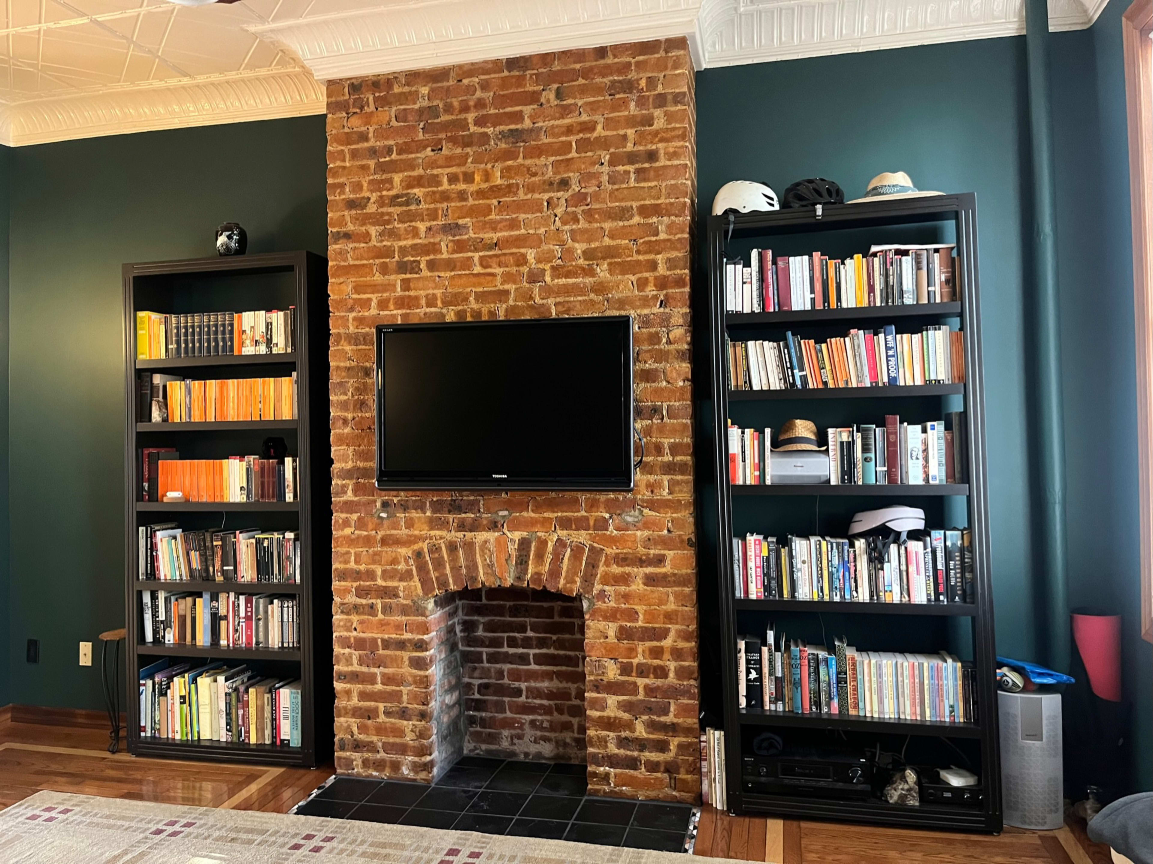 The image shows a living room featuring a brick fireplace with a television mounted above it, flanked by two tall black bookshelves filled with books.