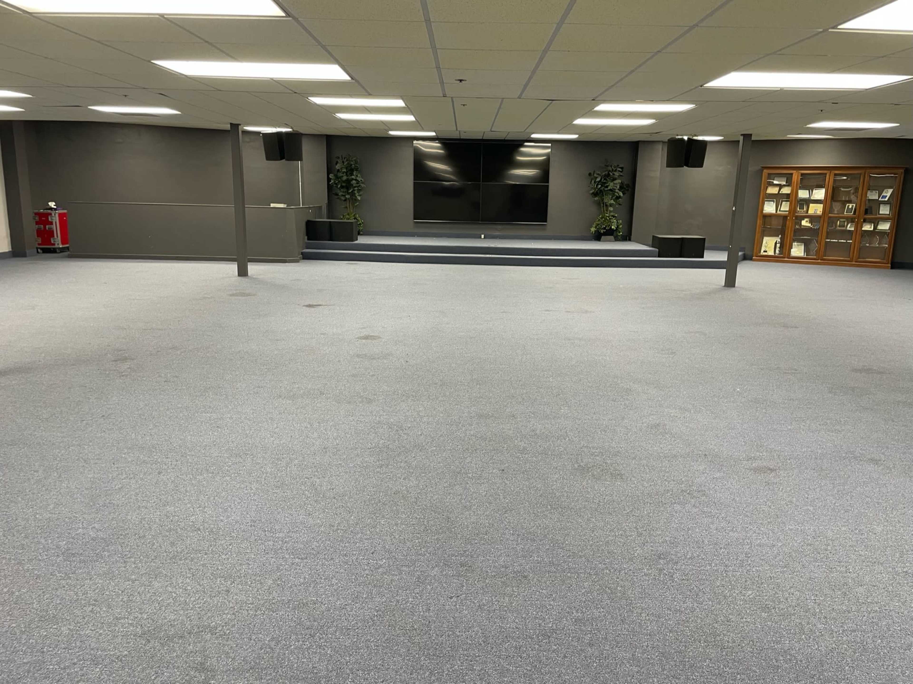 A spacious, empty room with grey carpet, a large screen at the front, and a display cabinet against the wall.