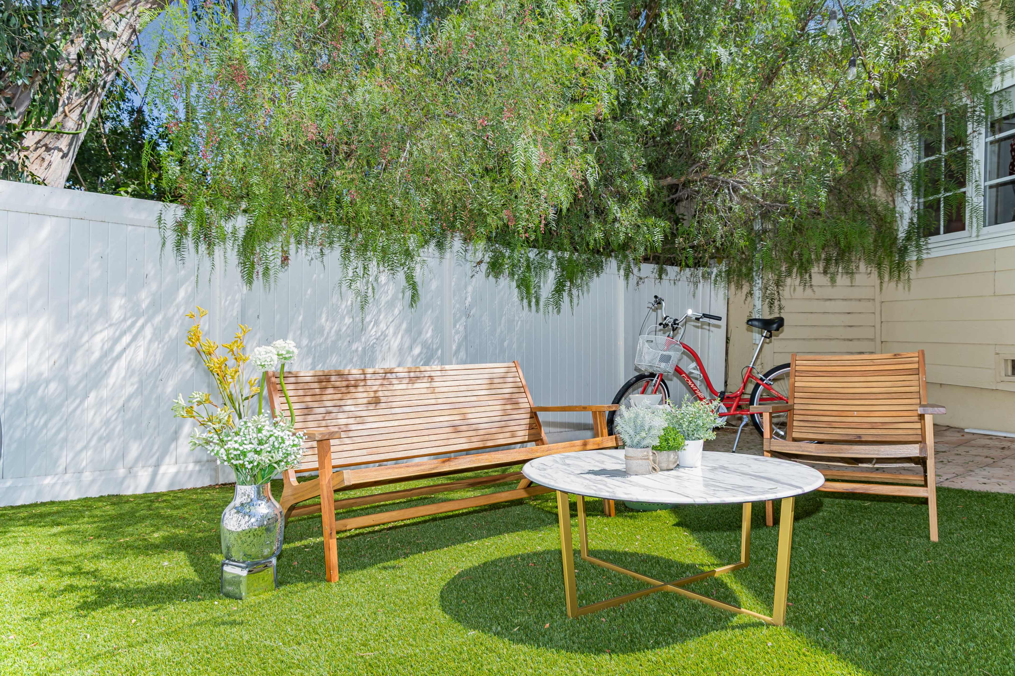 The image shows a small outdoor seating area with wooden furniture, a round marble table, and a bicycle leaning against a white fence amidst green grass.