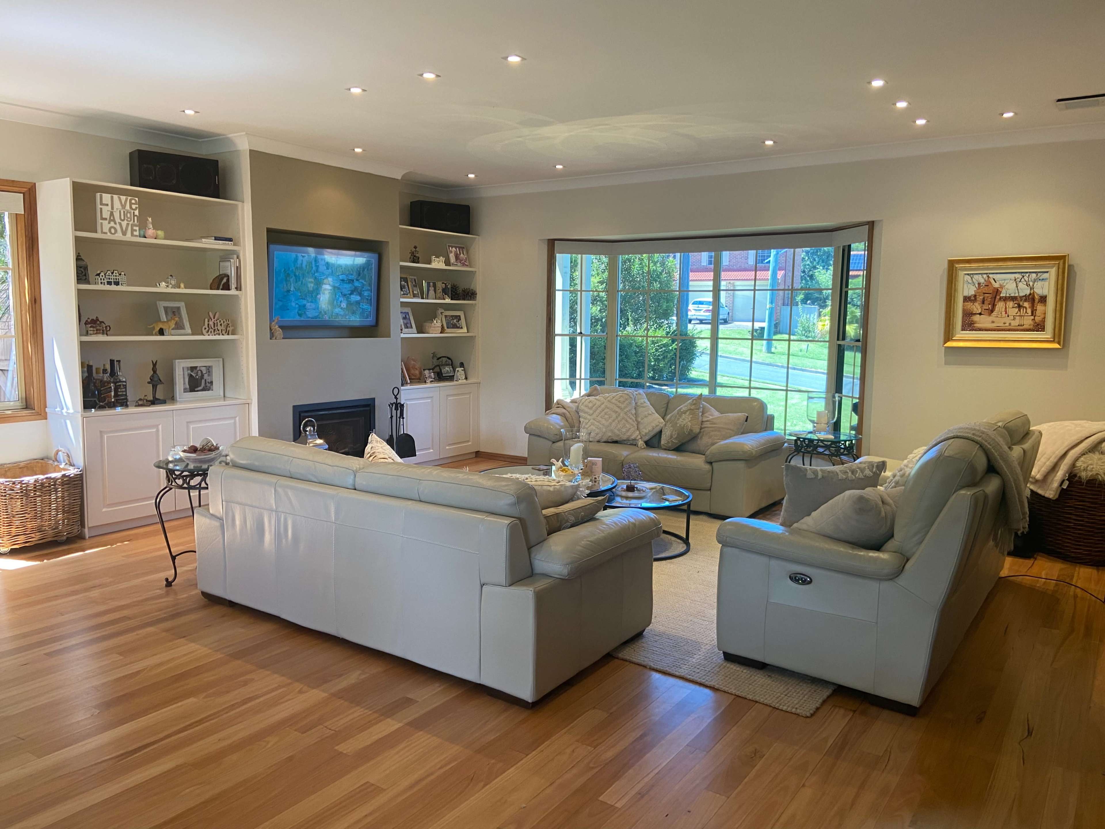 A spacious living room features light-colored leather sofas arranged around a coffee table, with a large window providing natural light and outdoor views.