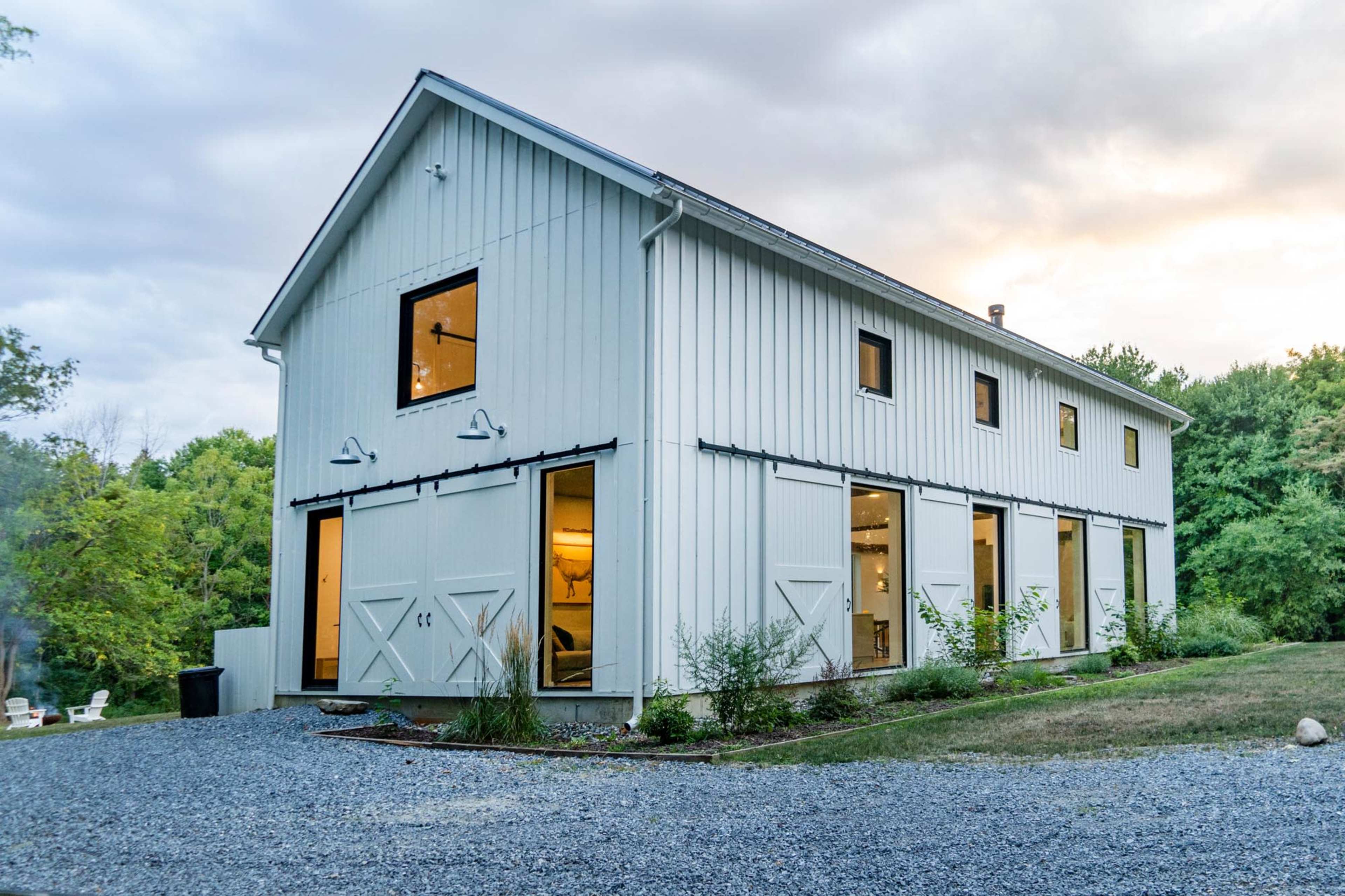 A white, two-story barn-style building with large windows and sliding doors is set on a gravel landscape surrounded by greenery.