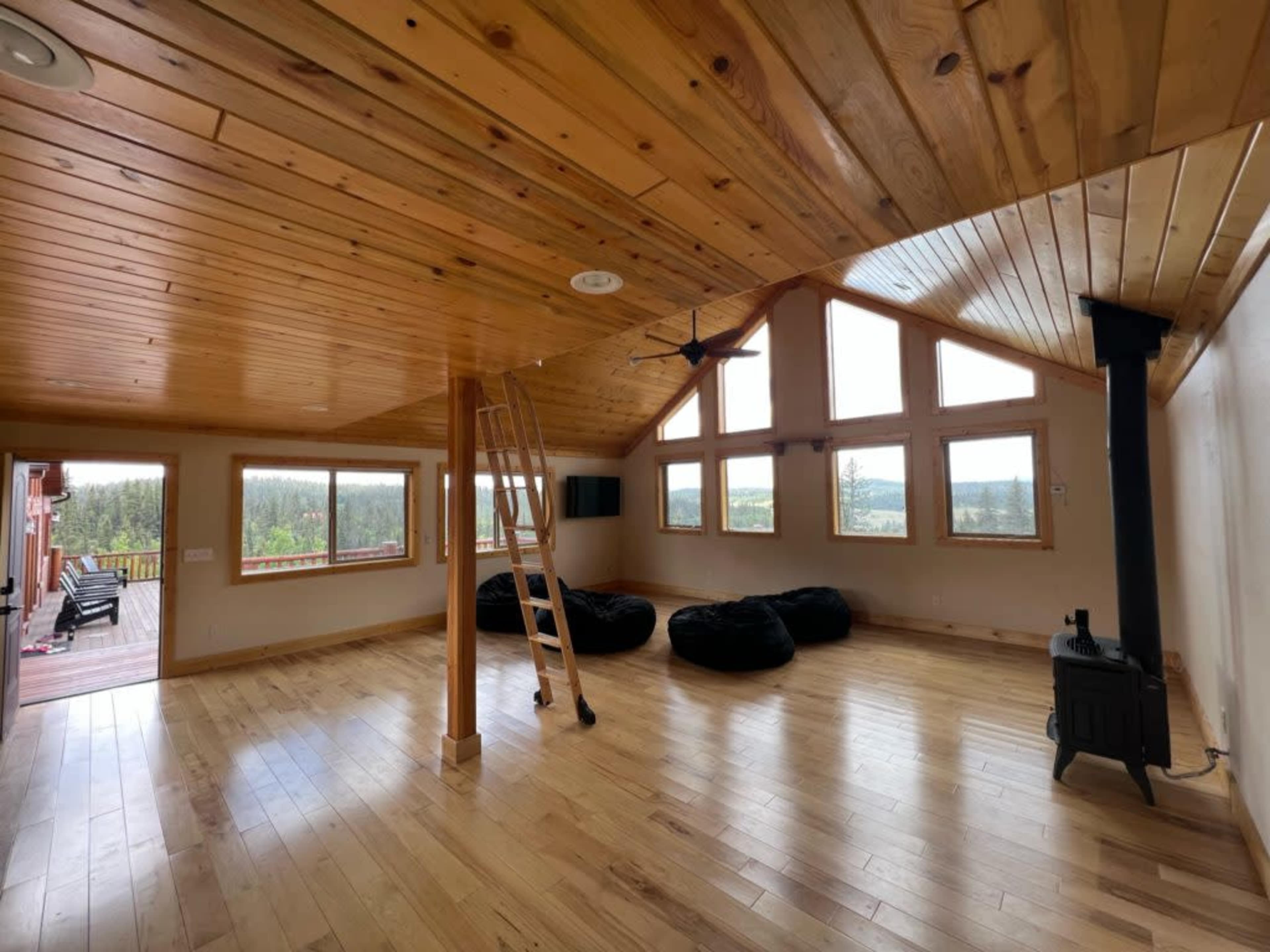The room features wooden walls and ceiling with large windows, a ladder, bean bag chairs, and a black wood stove in a spacious layout.
