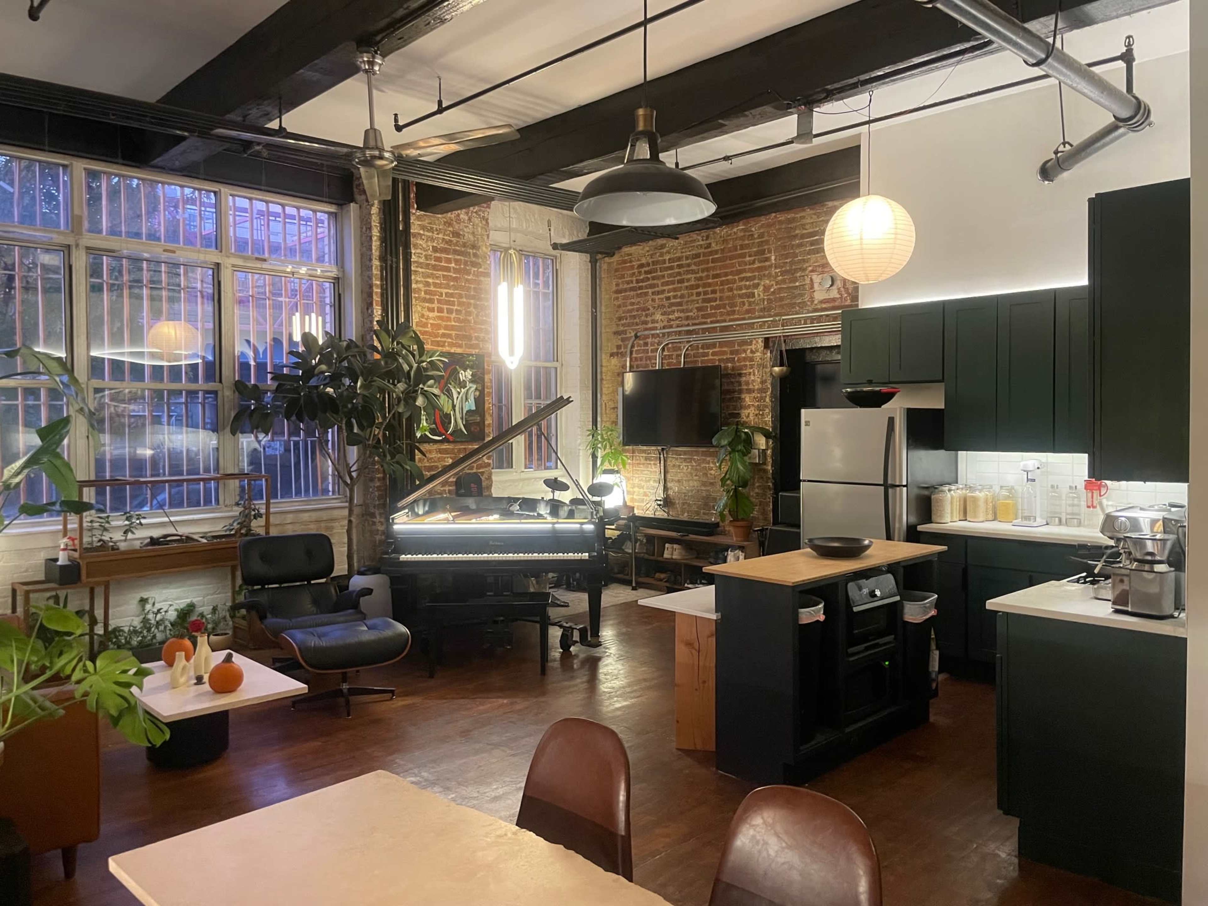 The image shows a spacious kitchen and living area featuring exposed brick walls, modern appliances, and a piano, with large windows allowing natural light to fill the space.