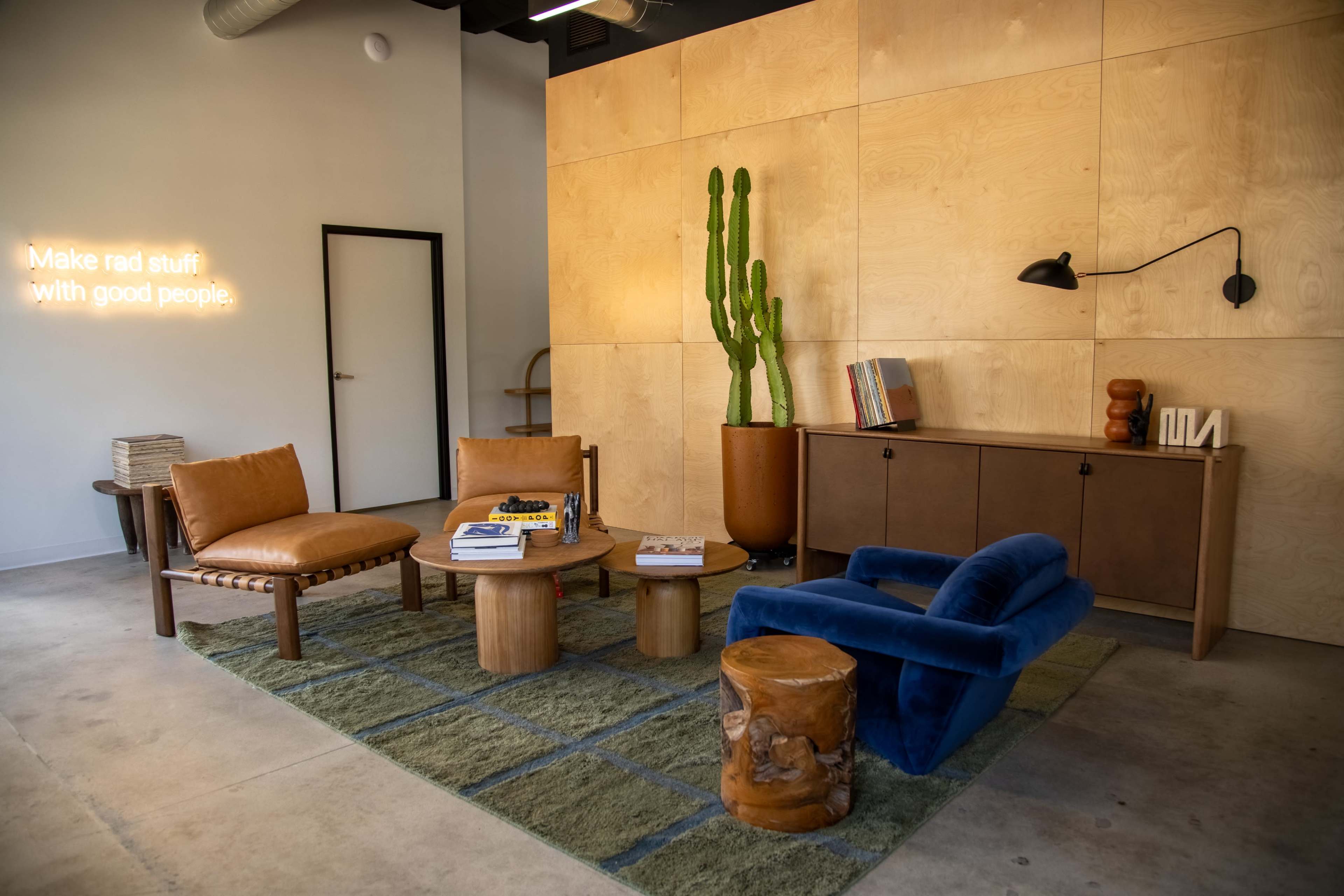 A modern lounge area featuring two brown armchairs, a blue sofa, a round wooden coffee table, and a green rug, with a cactus and wooden storage cabinet in the background.