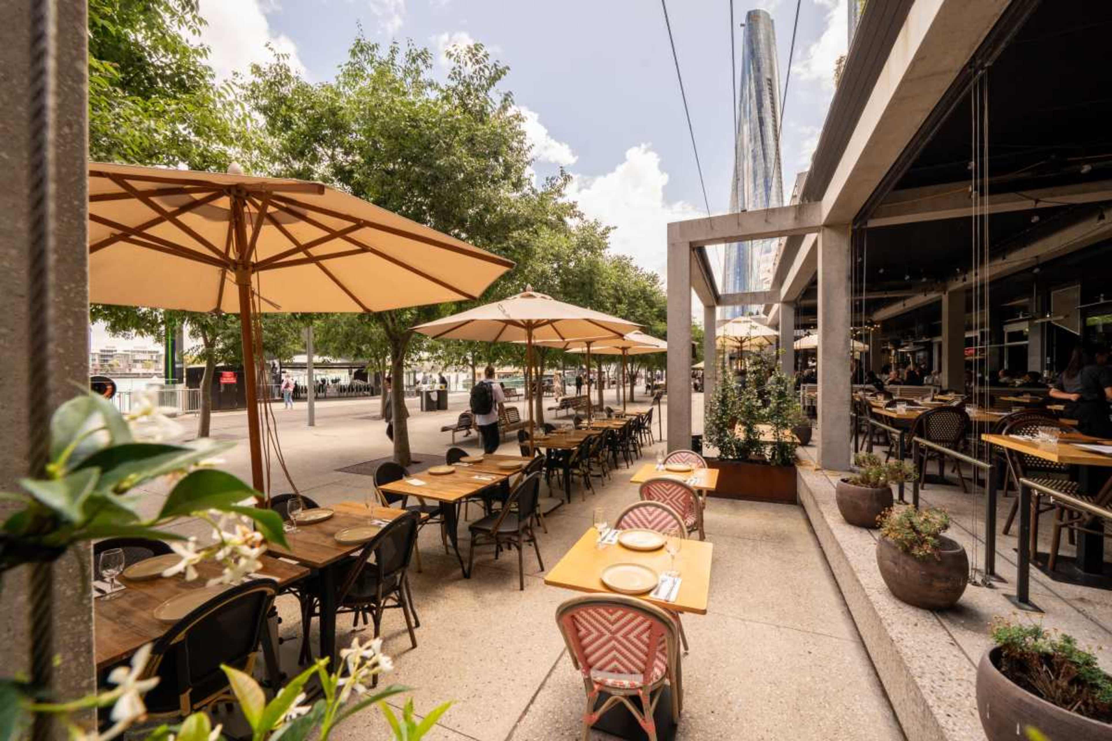 An outdoor dining area of a restaurant lined with tables and umbrellas along a sidewalk, surrounded by trees and a modern building in the background.