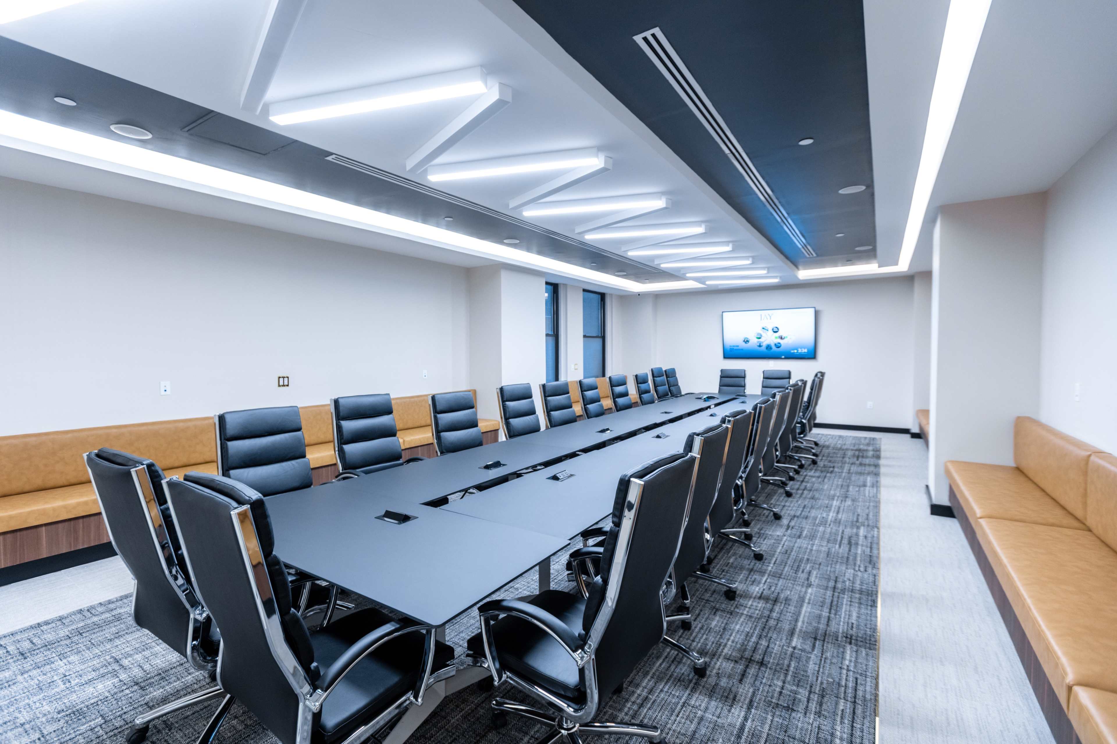 A long conference table with black chairs is arranged in a modern meeting room featuring a large screen and soft seating along one wall.