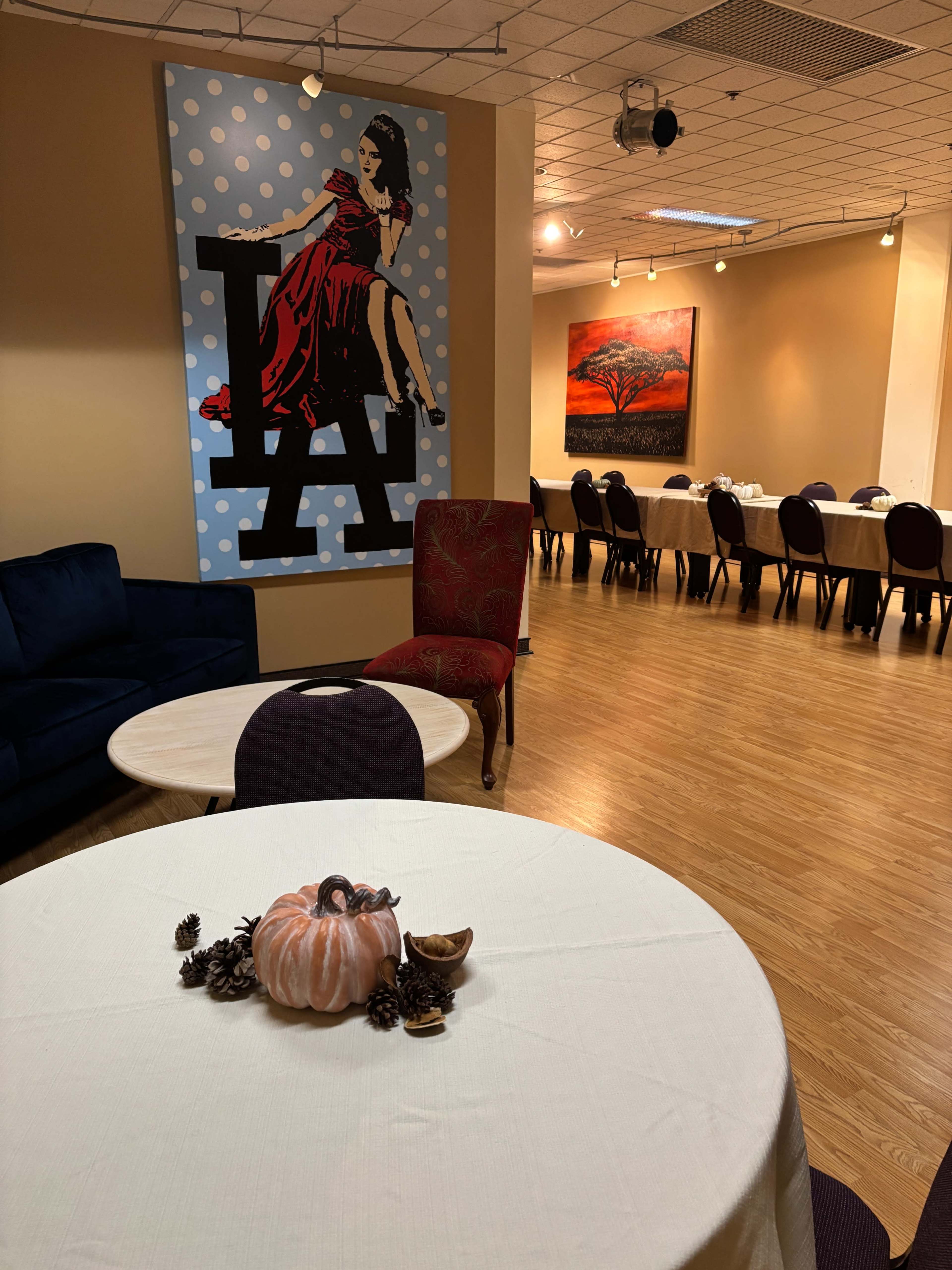 The image shows a room with a blue polka-dotted wall featuring a large artwork, a seating area with a blue couch and a chair, and a table set with a decorative pumpkin and pinecones in the foreground.