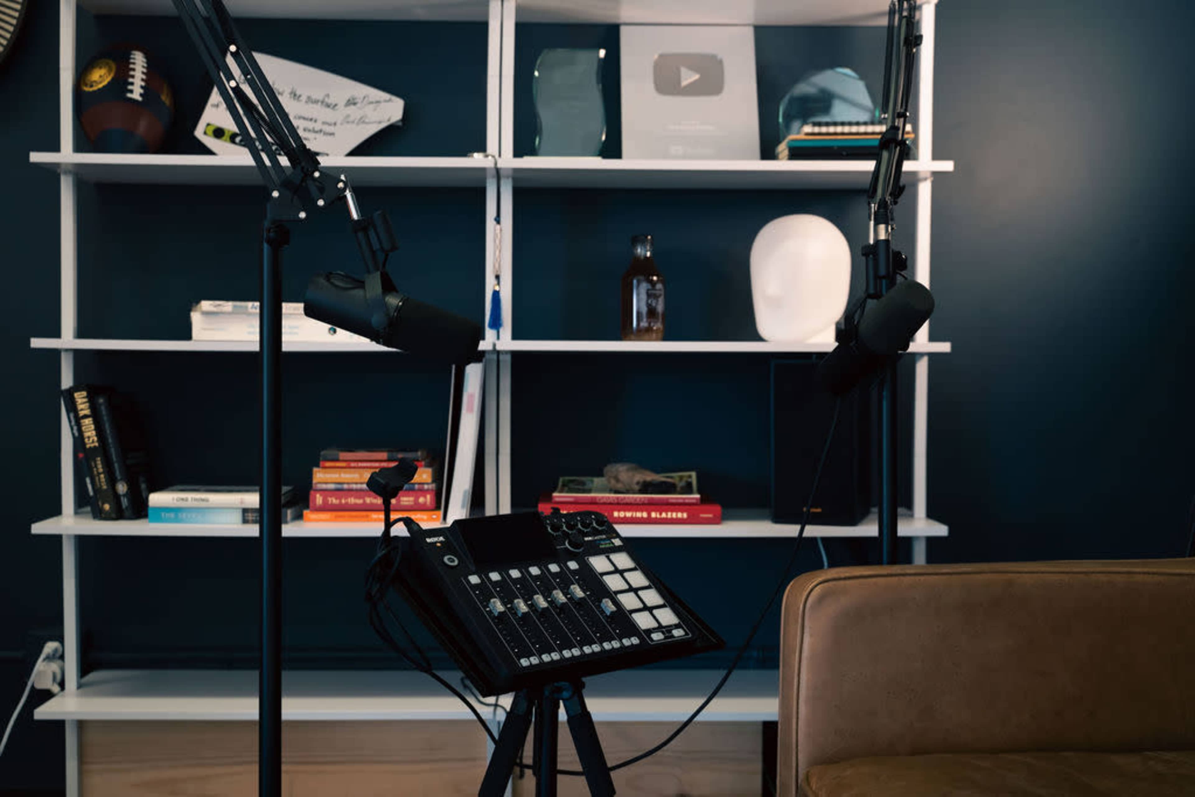 The image shows a well-organized podcasting setup with two microphones on adjustable stands, an audio mixer, and a bookshelf displaying various items and awards.