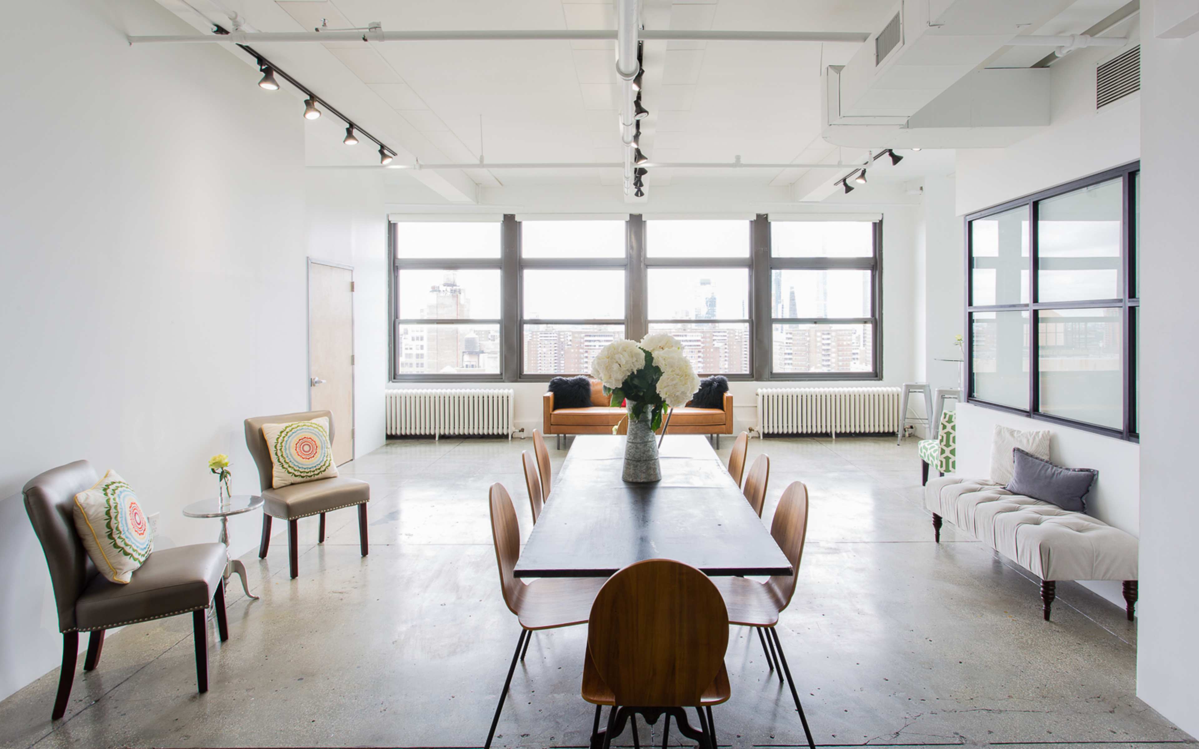 A modern dining area with a long table surrounded by chairs, large windows offering a city view, and a mix of seating arrangements against the walls.