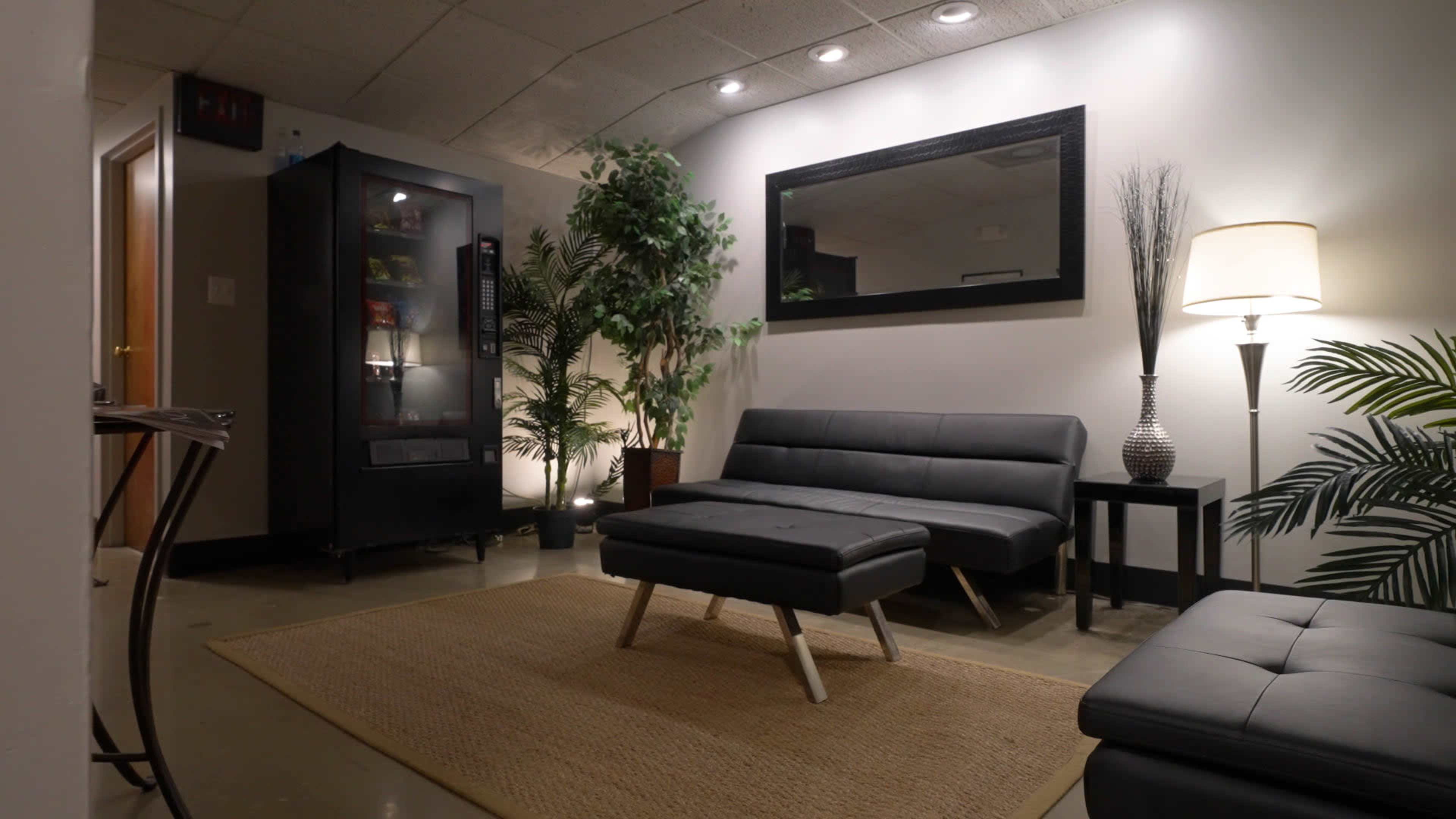 A modern lounge area features a black sofa, an ottoman, a decorative plant, and a tall lamp, all arranged beneath soft lighting.