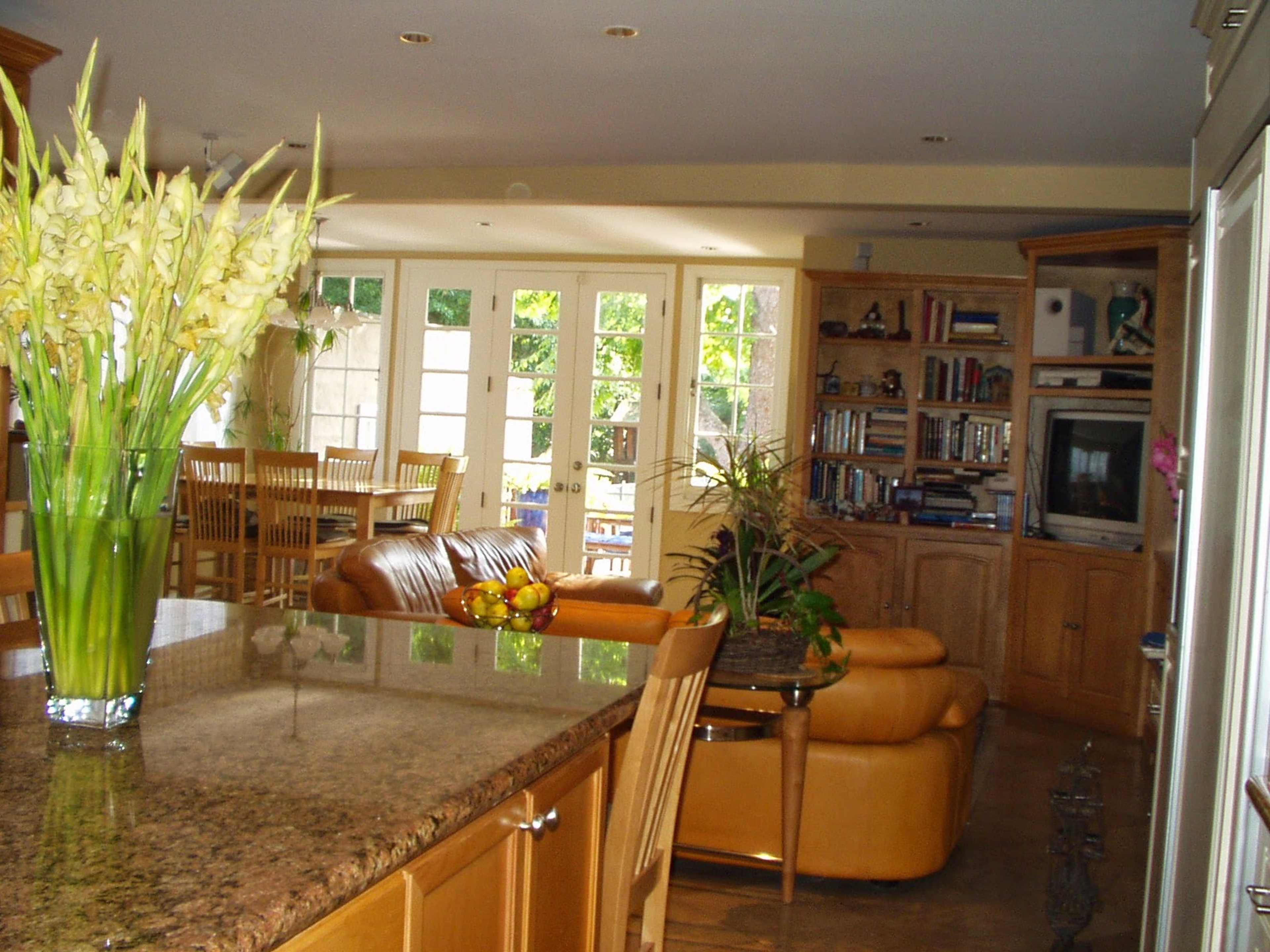 A spacious kitchen features a granite countertop, a large floral arrangement, and connects to a living area with bookshelves and large windows.