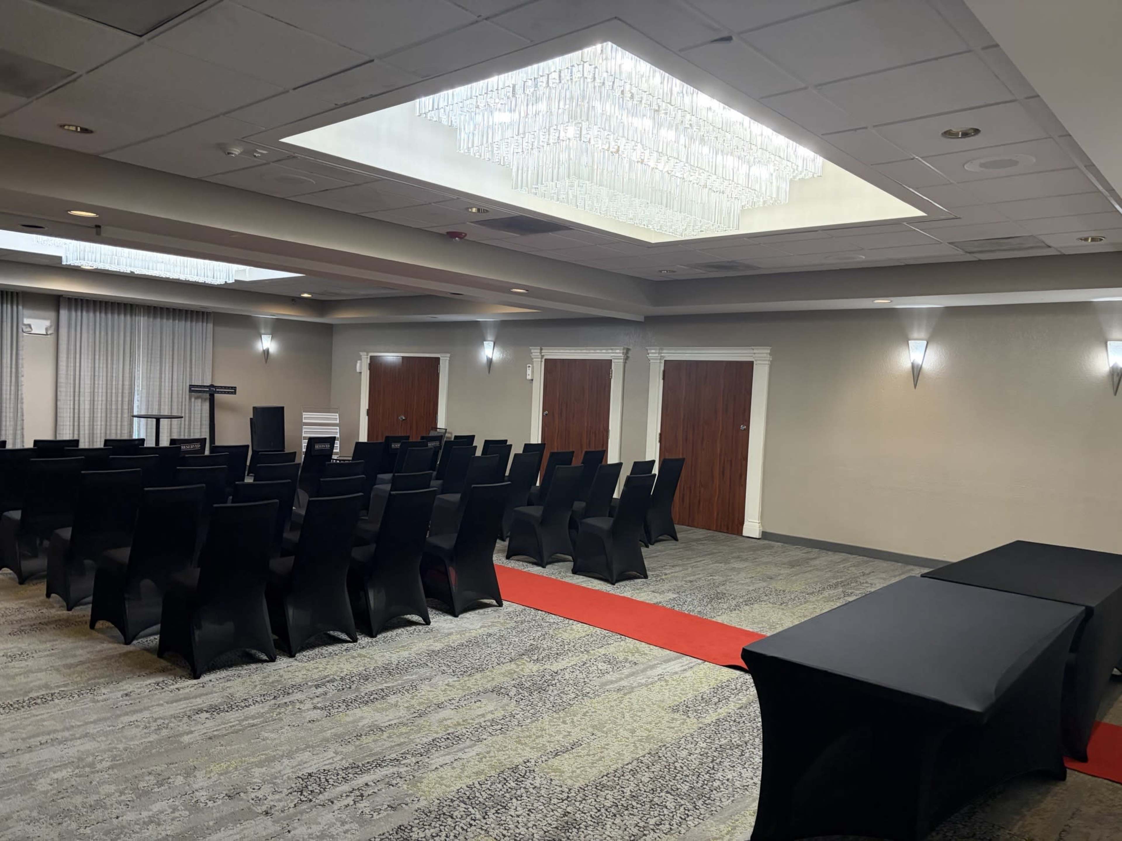 A conference room is set up with rows of black chairs facing a presentation area, featuring a red carpet and tables along one side.