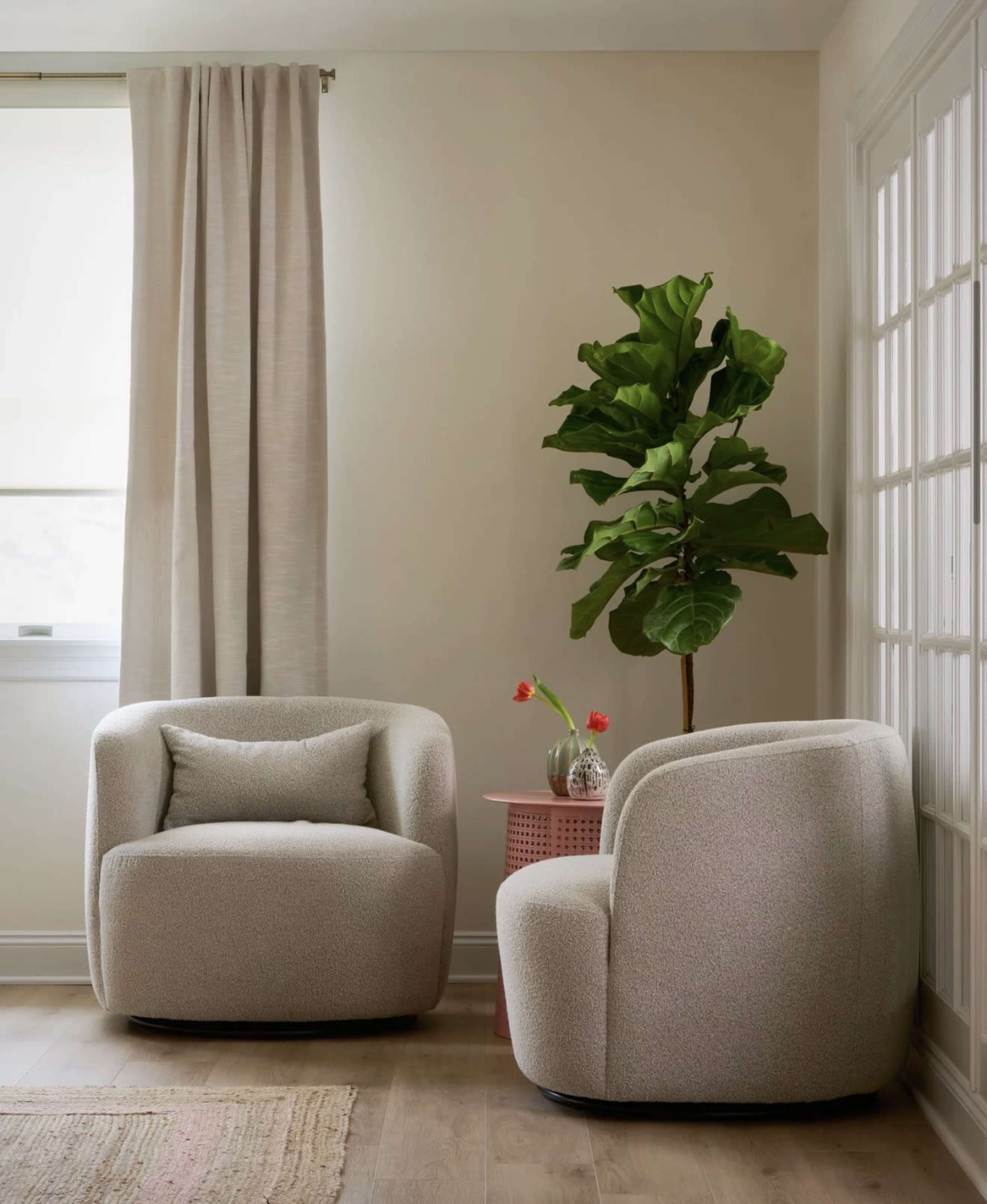The image shows two cozy, light-colored chairs positioned next to a small round table with a vase of flowers, against a backdrop of a large window and a potted plant.