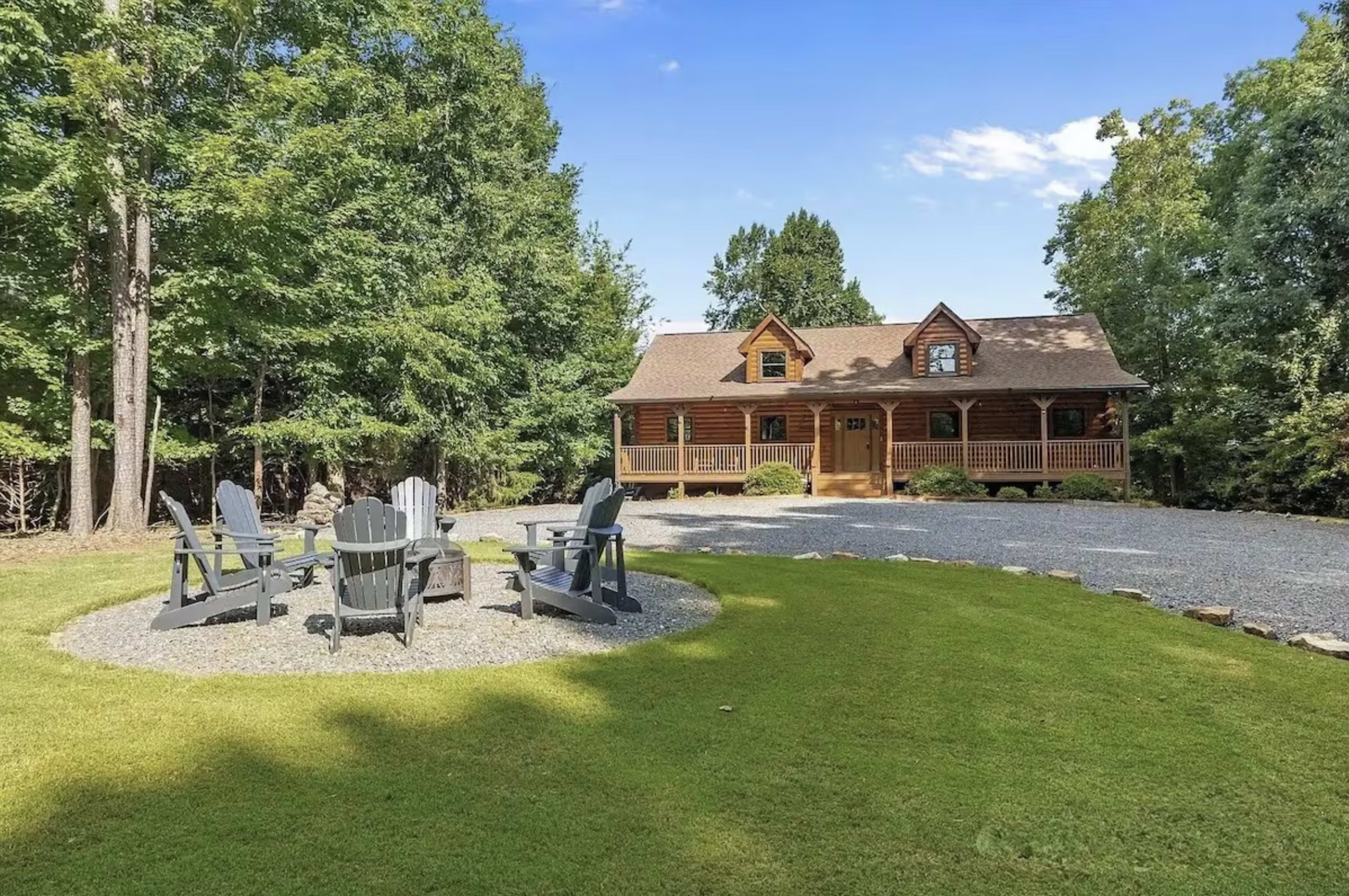 A log cabin with a gravel driveway is surrounded by trees and features a circular arrangement of Adirondack chairs on a grassy area in front.