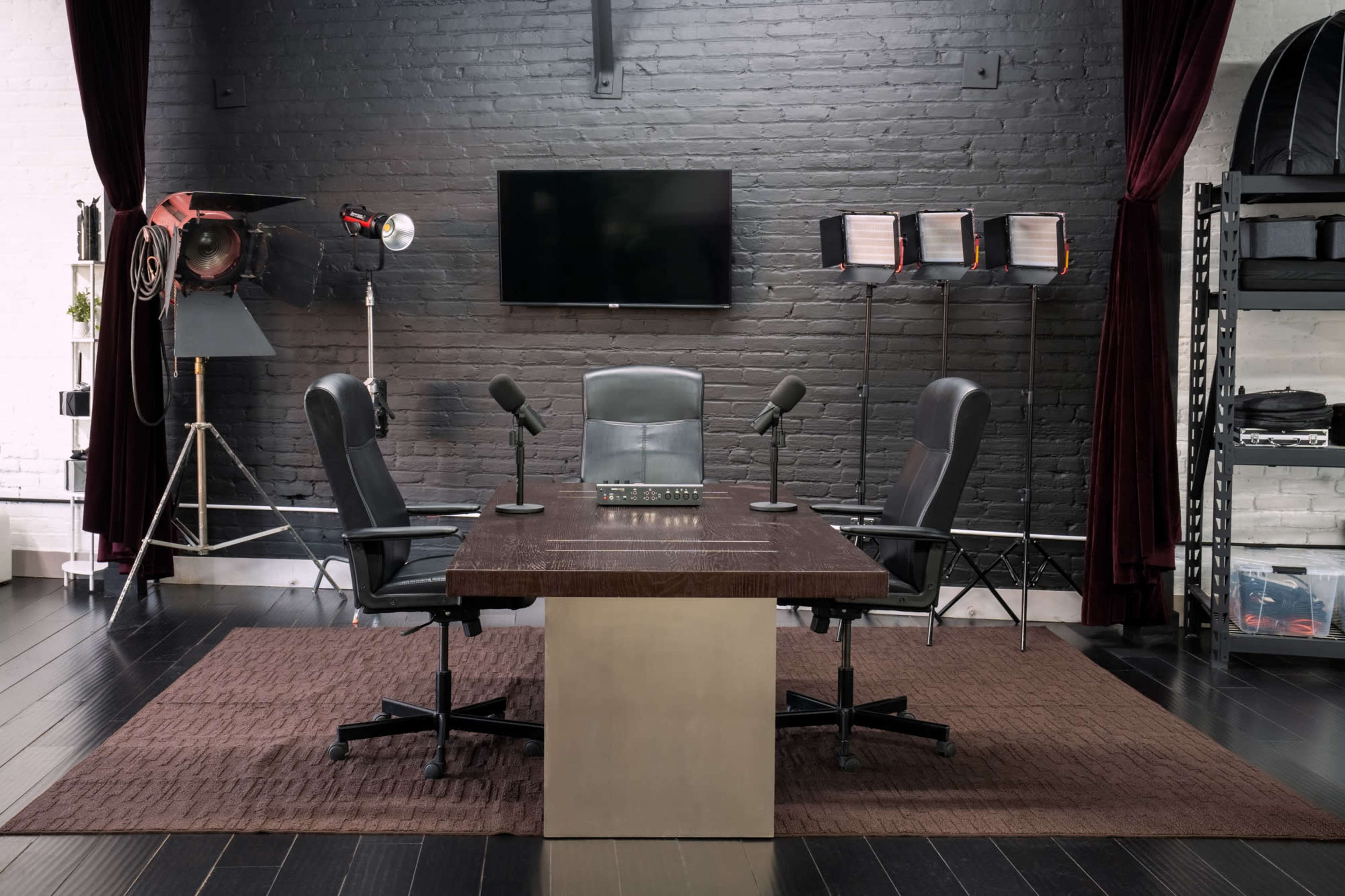 The image shows a dimly lit conference room with a large table, two office chairs, and a television mounted on a brick wall, accompanied by studio lighting equipment in the background.