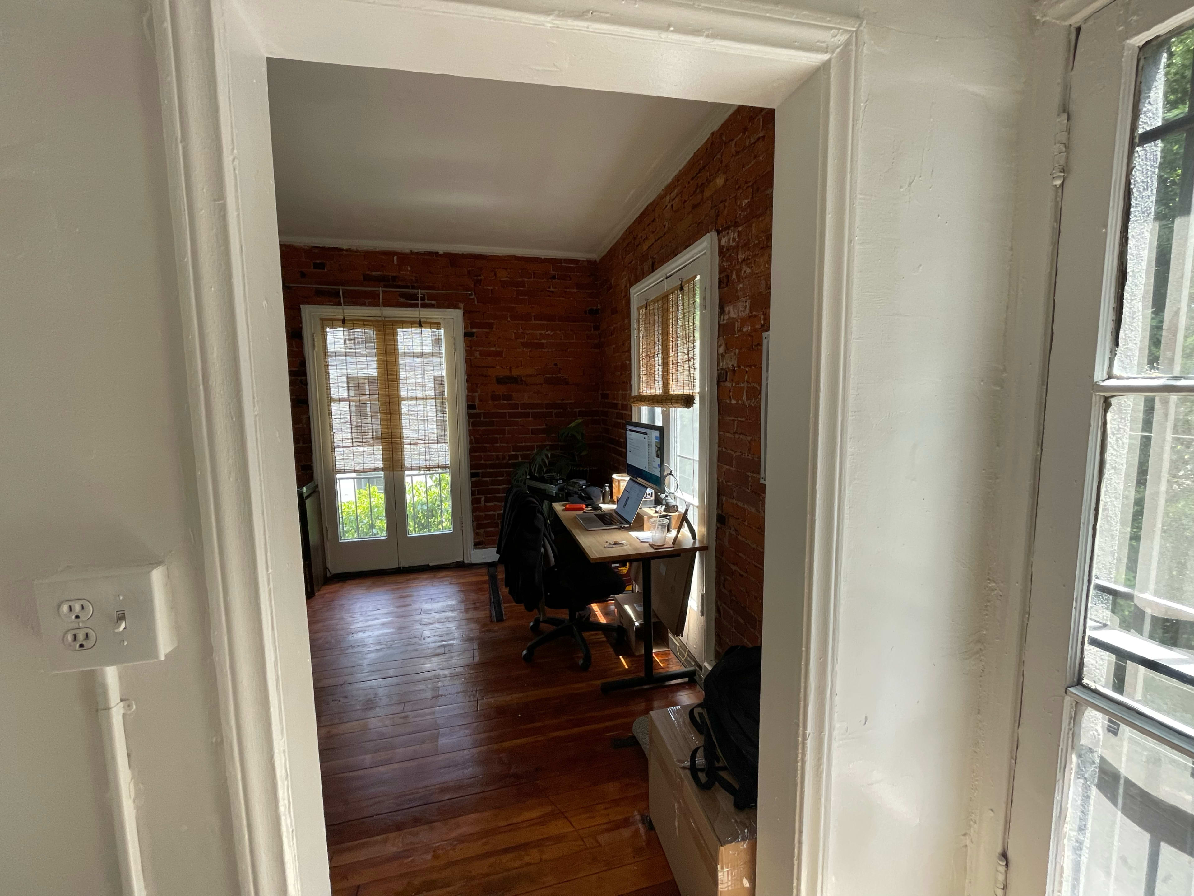 The image shows a room with exposed brick walls, a desk with a computer, and an adjacent window with partially opened blinds.
