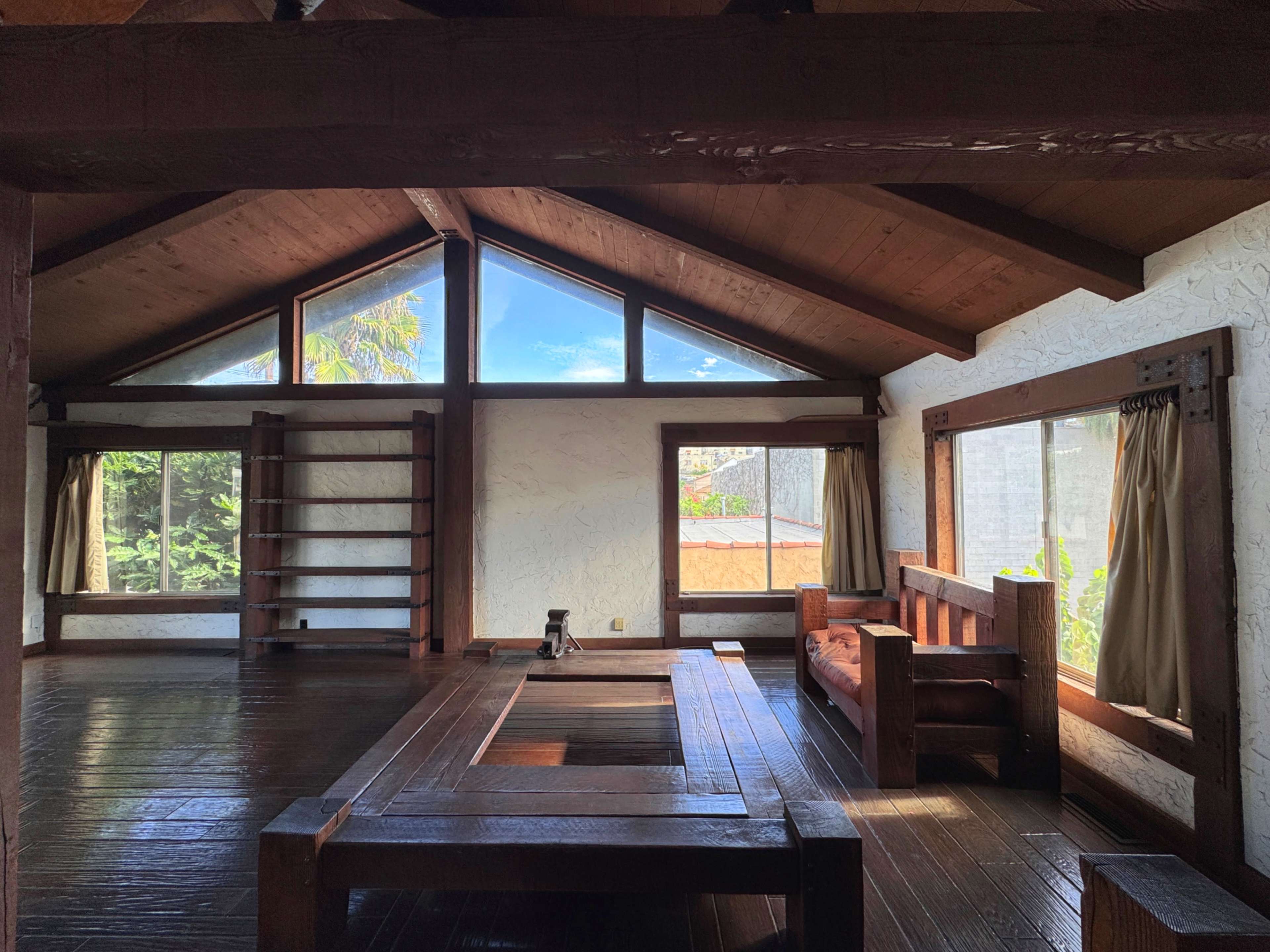 The interior of a wooden cabin features high ceilings, large windows with natural light, and minimalist furnishings including a wooden table and bench.