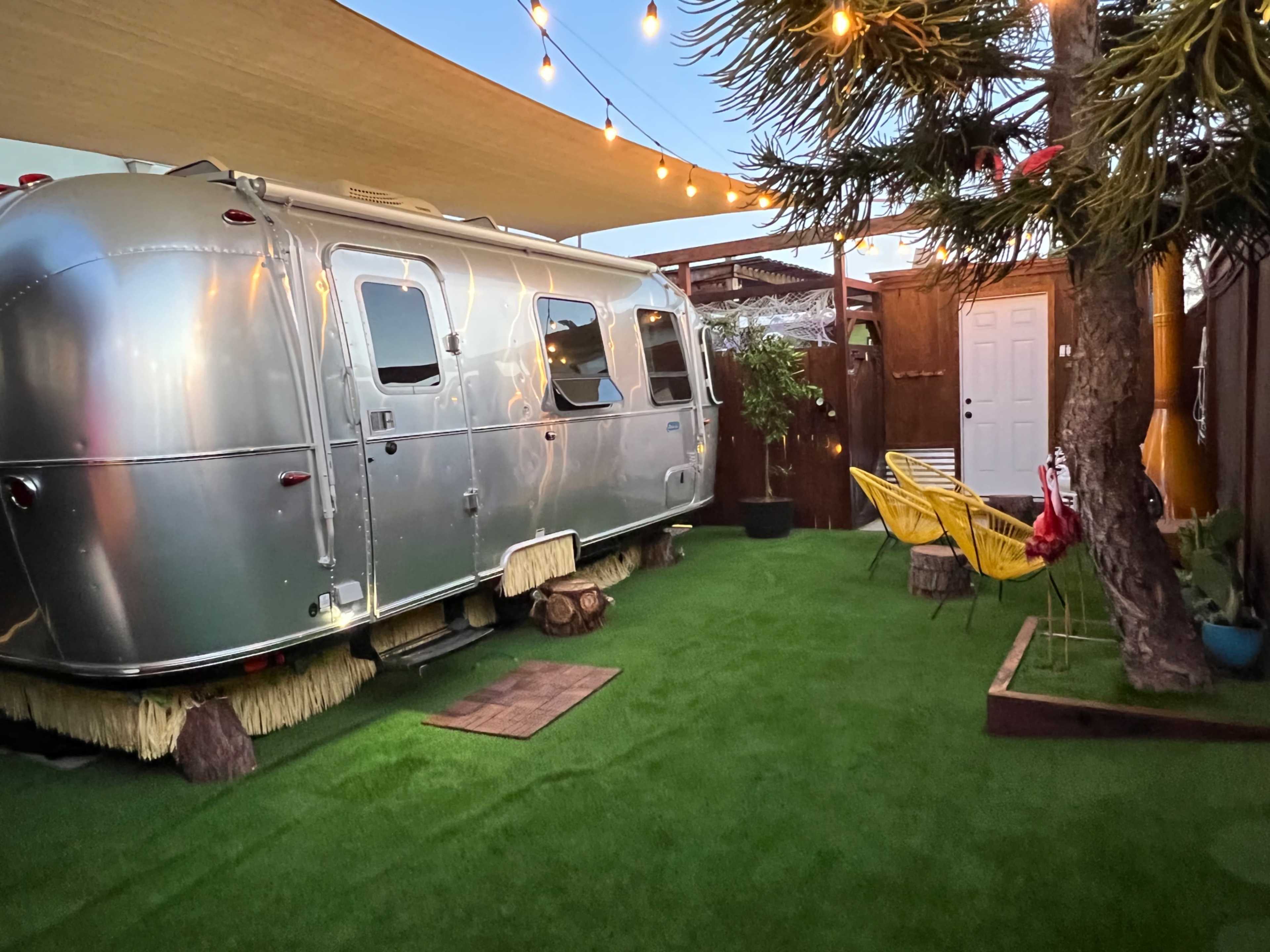 A silver Airstream trailer is parked on a grassy area surrounded by string lights and potted plants.