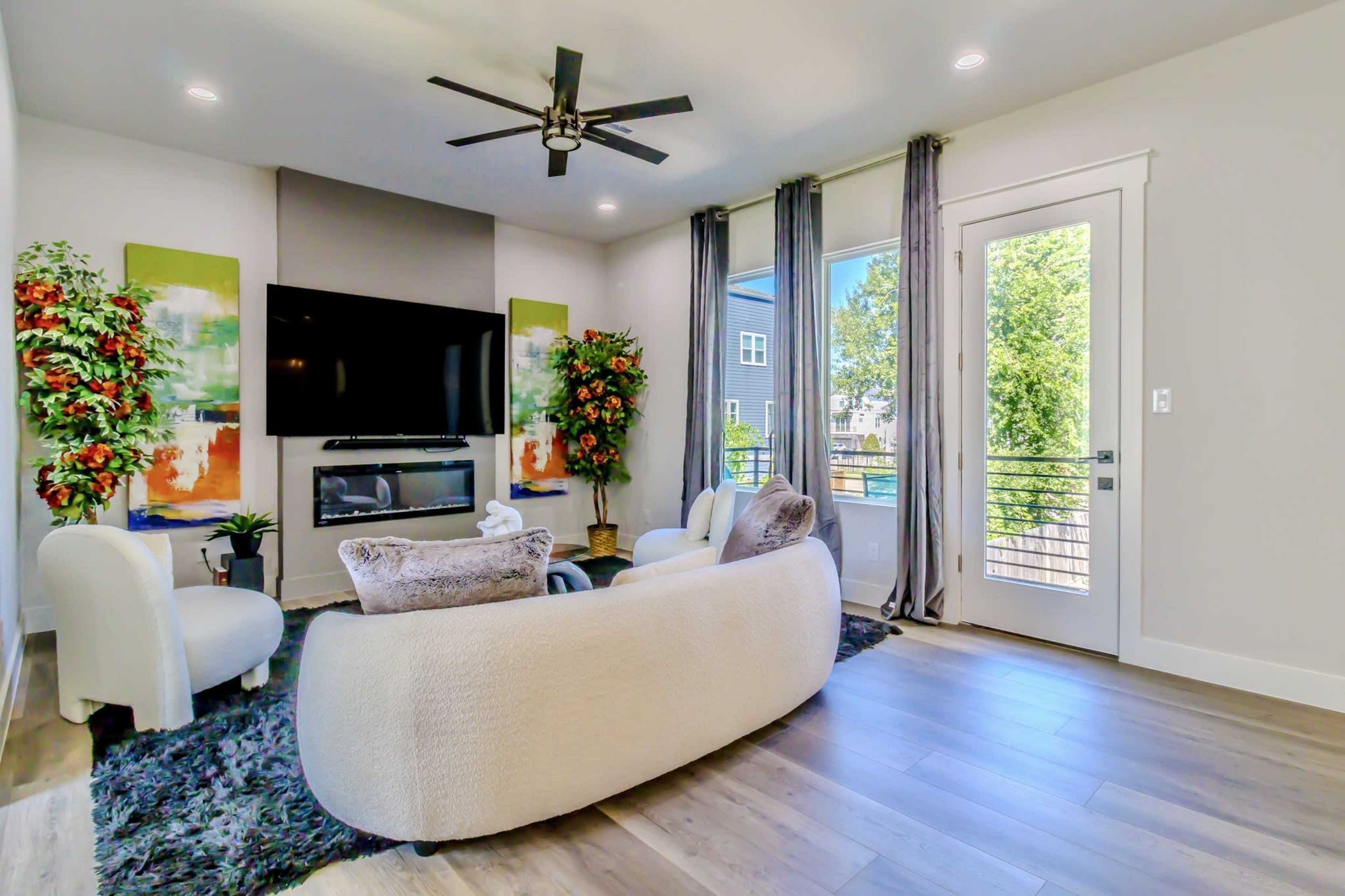 A modern living room features a large television on a wall with two decorative plants, a round rug, and a light-colored sofa set near a balcony door that overlooks greenery.