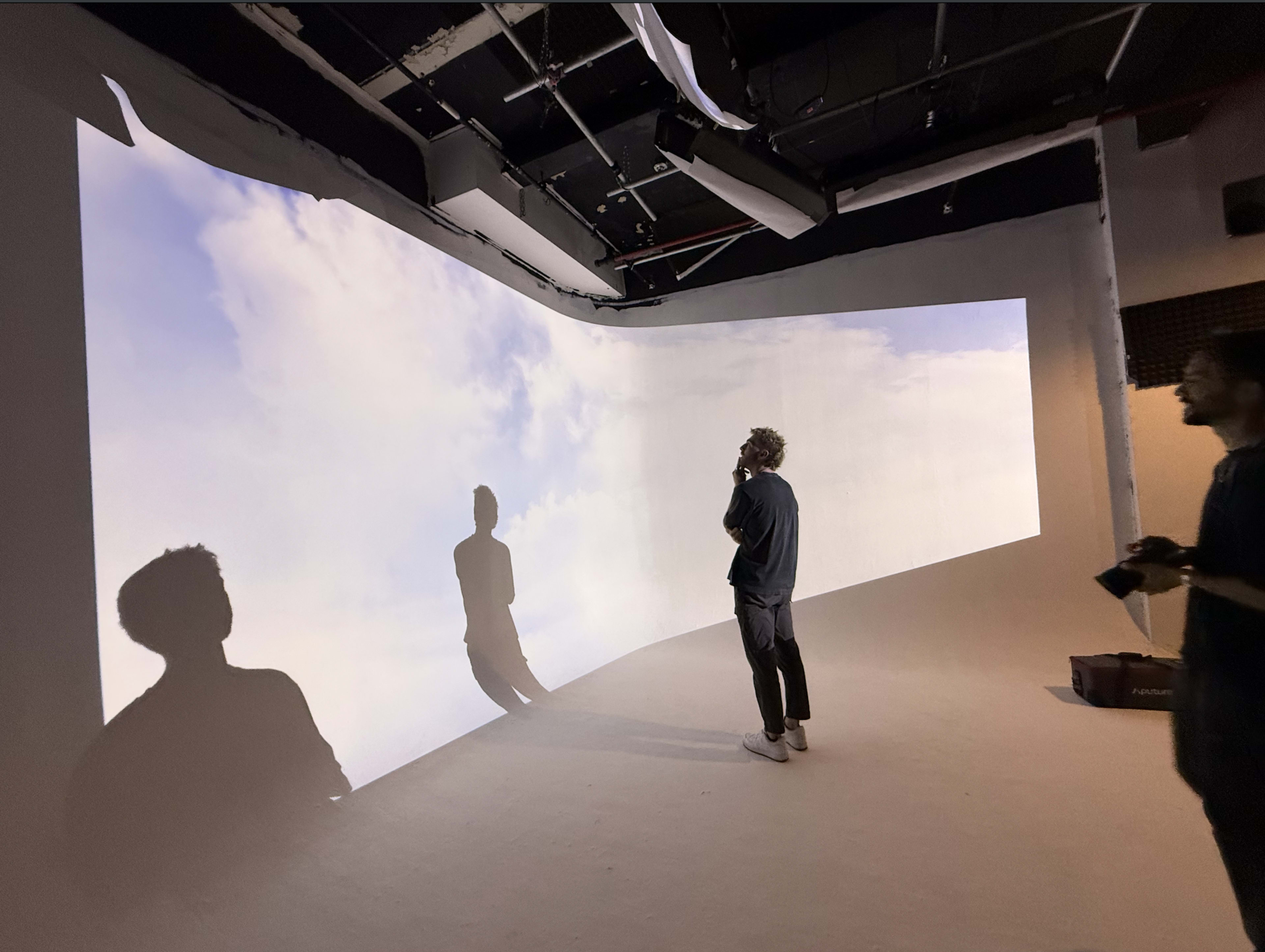 The image shows two people standing in a room with a curved projection of clouds on the wall, casting their shadows on the floor.