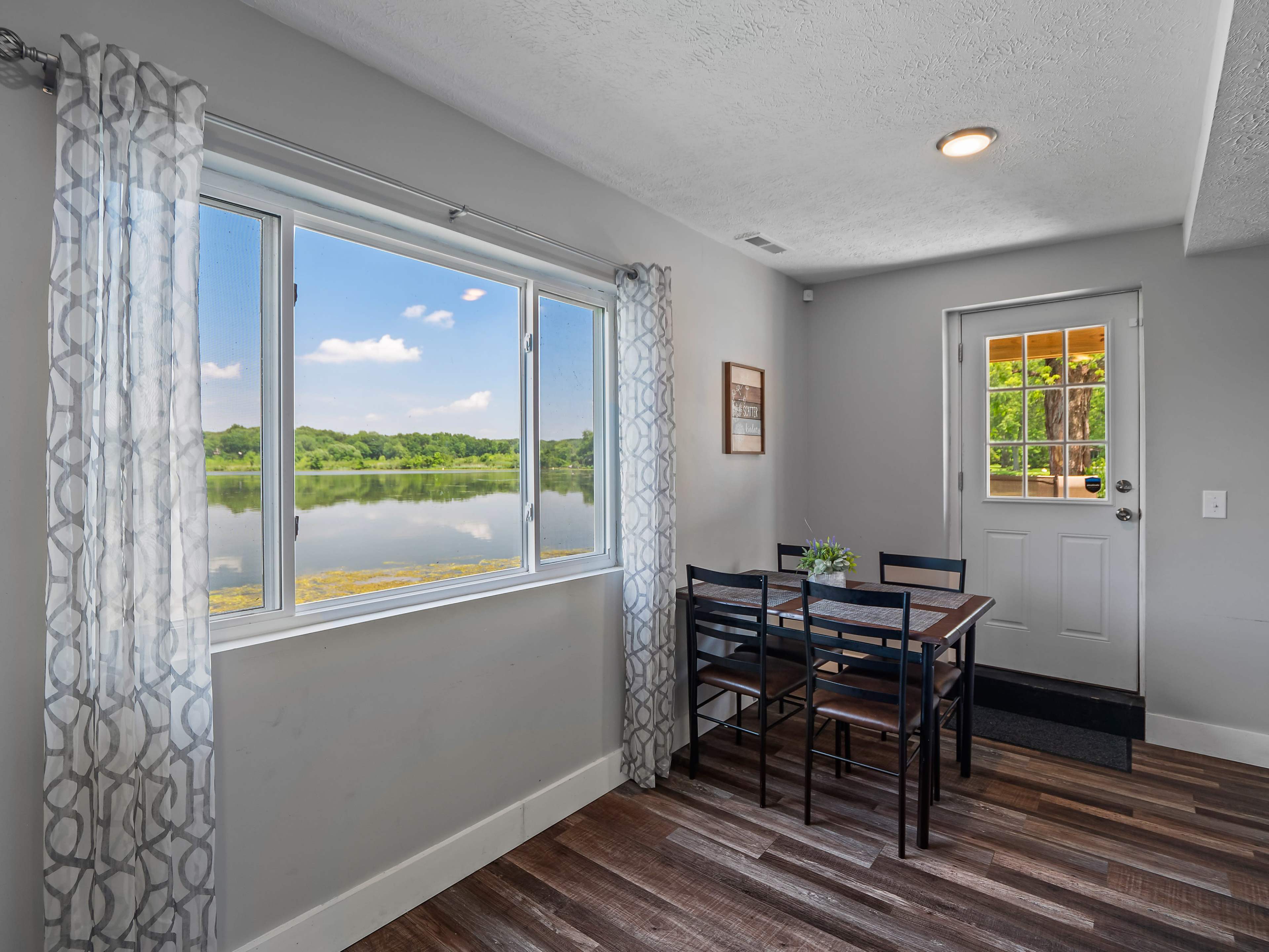 A dining area with a table and chairs near a large window overlooking a calm body of water and greenery outside.