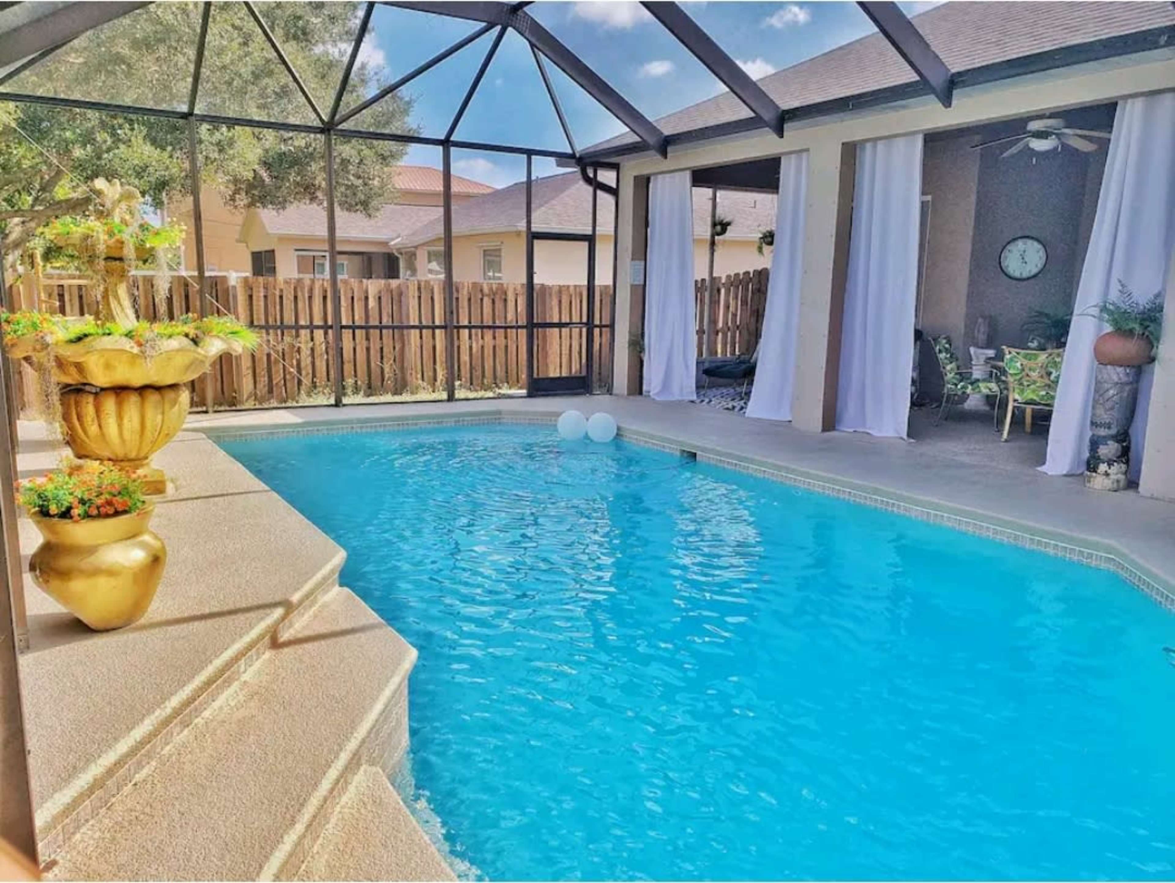 A screened-in pool area featuring a clear blue pool, a decorative planter, and seating arrangements with curtains on either side.