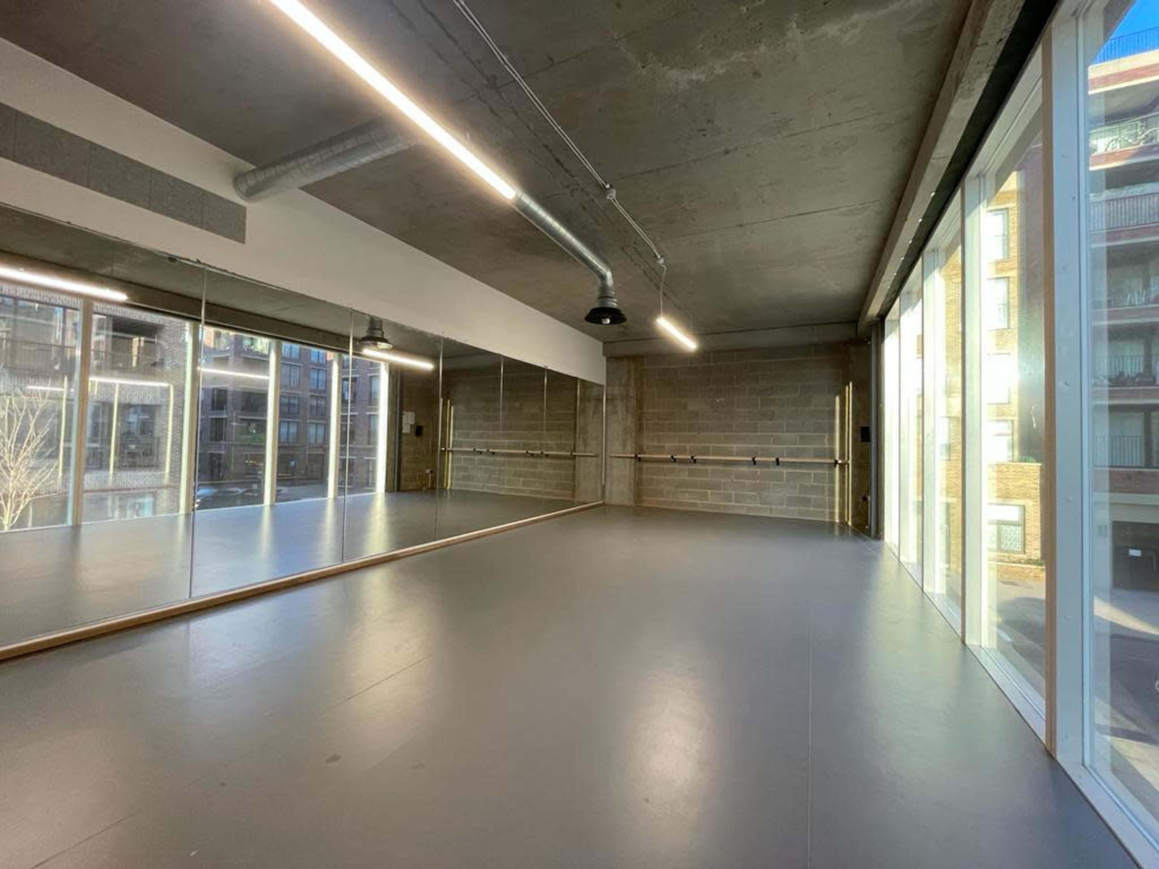 A spacious dance studio with large mirrors, a barre along one wall, and floor-to-ceiling windows providing natural light.