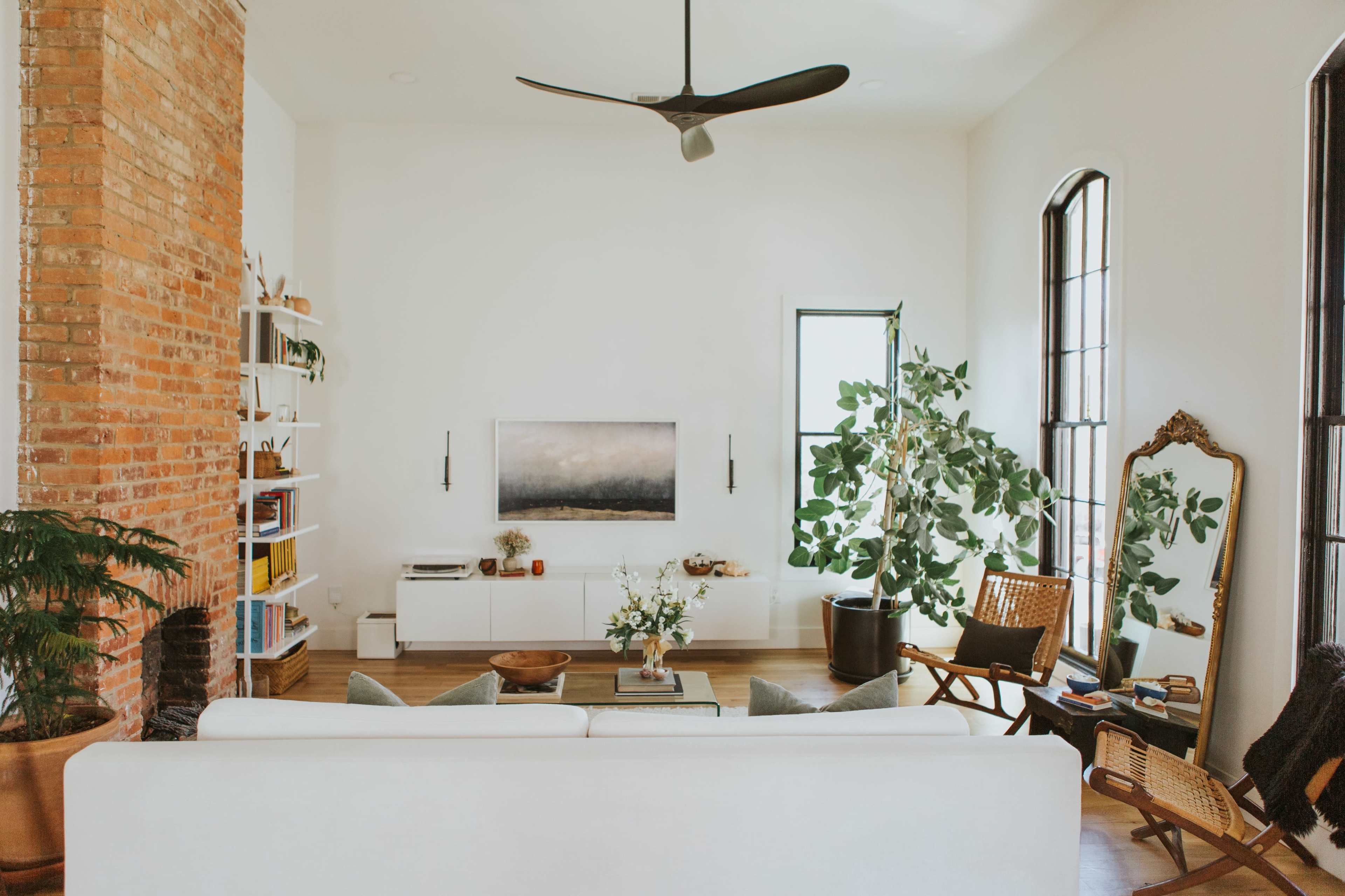 A modern living room features a white sofa, a large potted plant, a television mounted on the wall, and a wooden coffee table surrounded by various seating options.