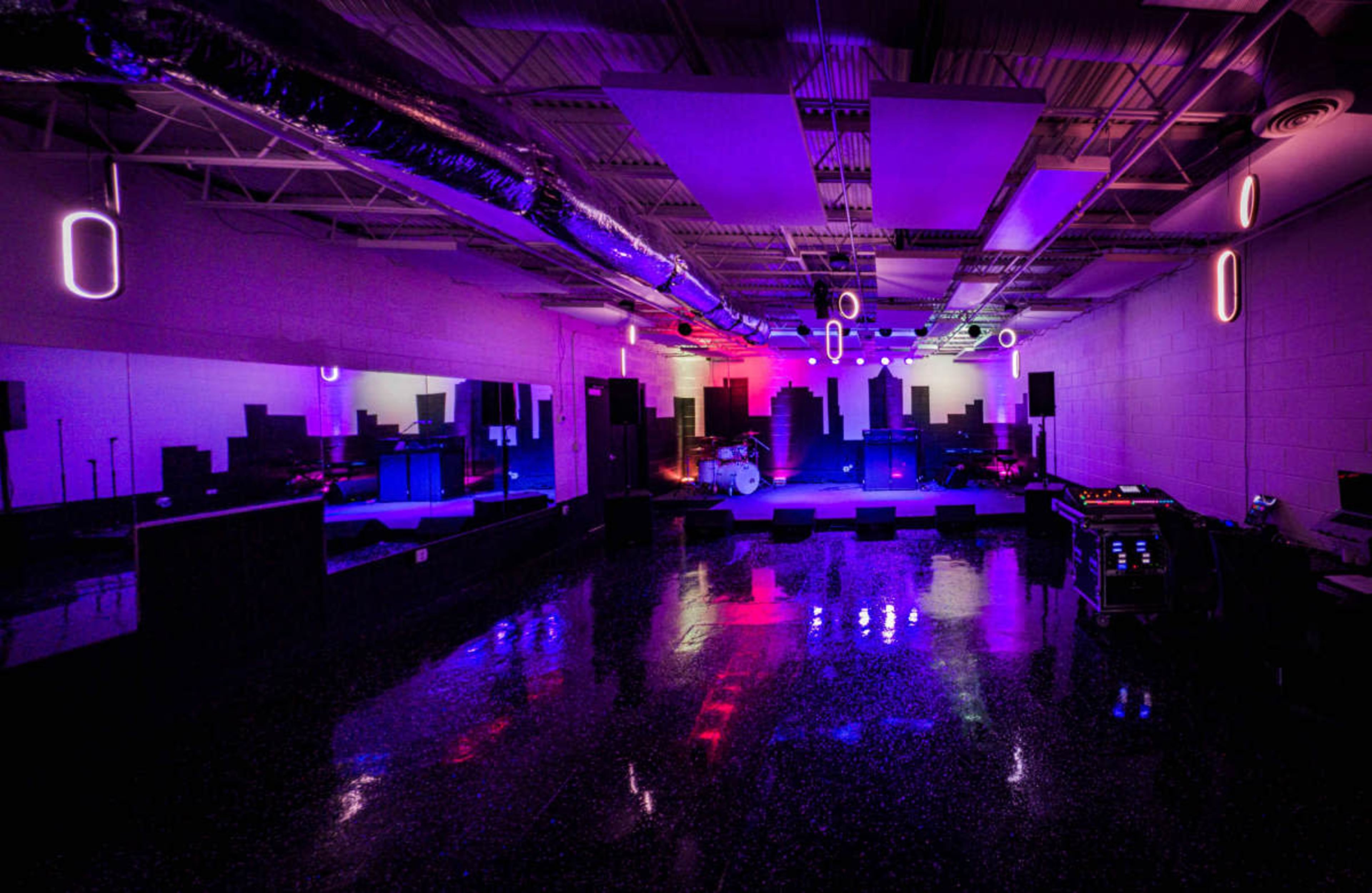 The image shows a music venue with a polished floor, stage area, and colorful lighting, featuring sound equipment and reflective surfaces.