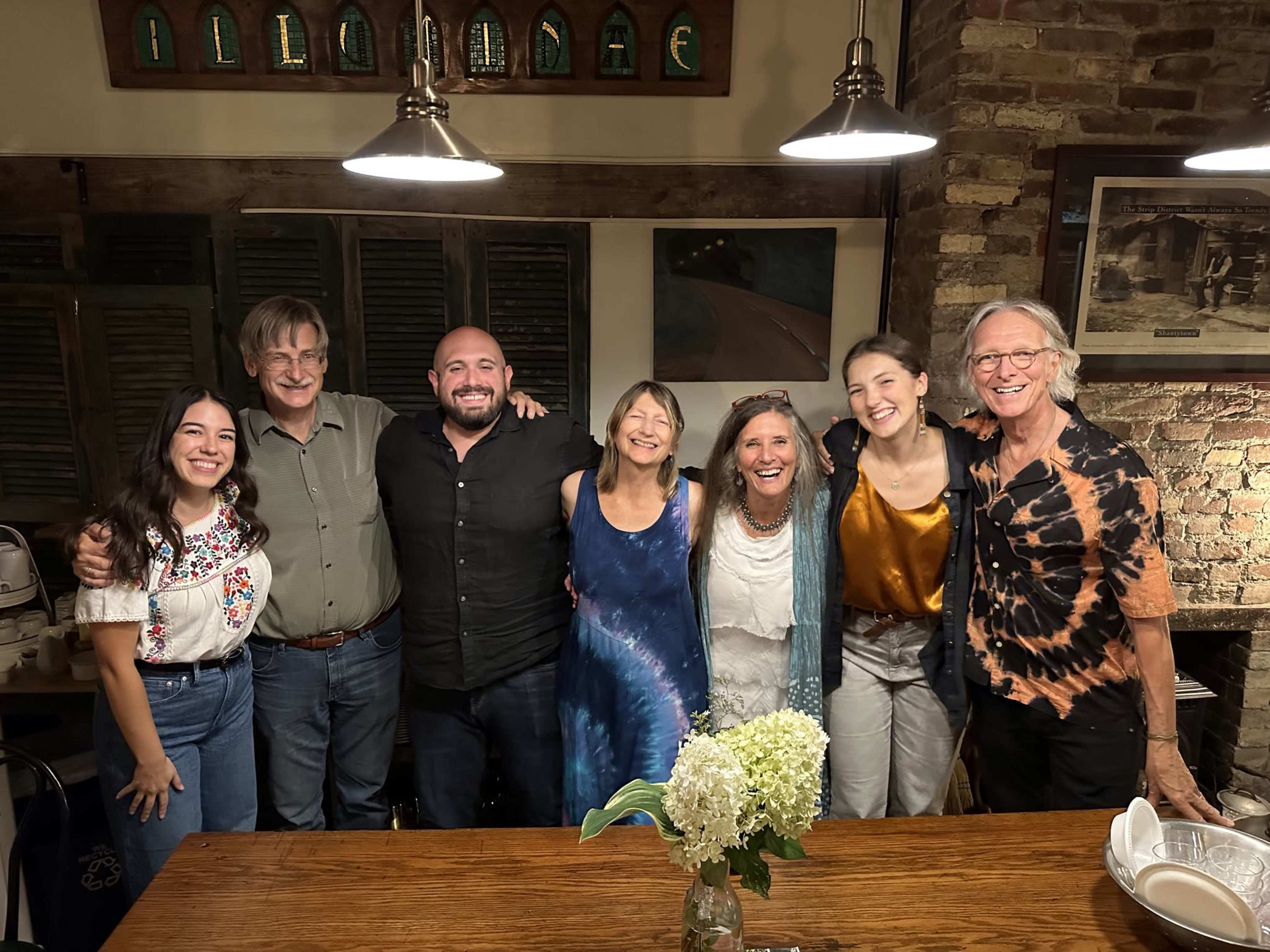 A group of seven people stands together in a rustic dining area, smiling and posing for the camera.