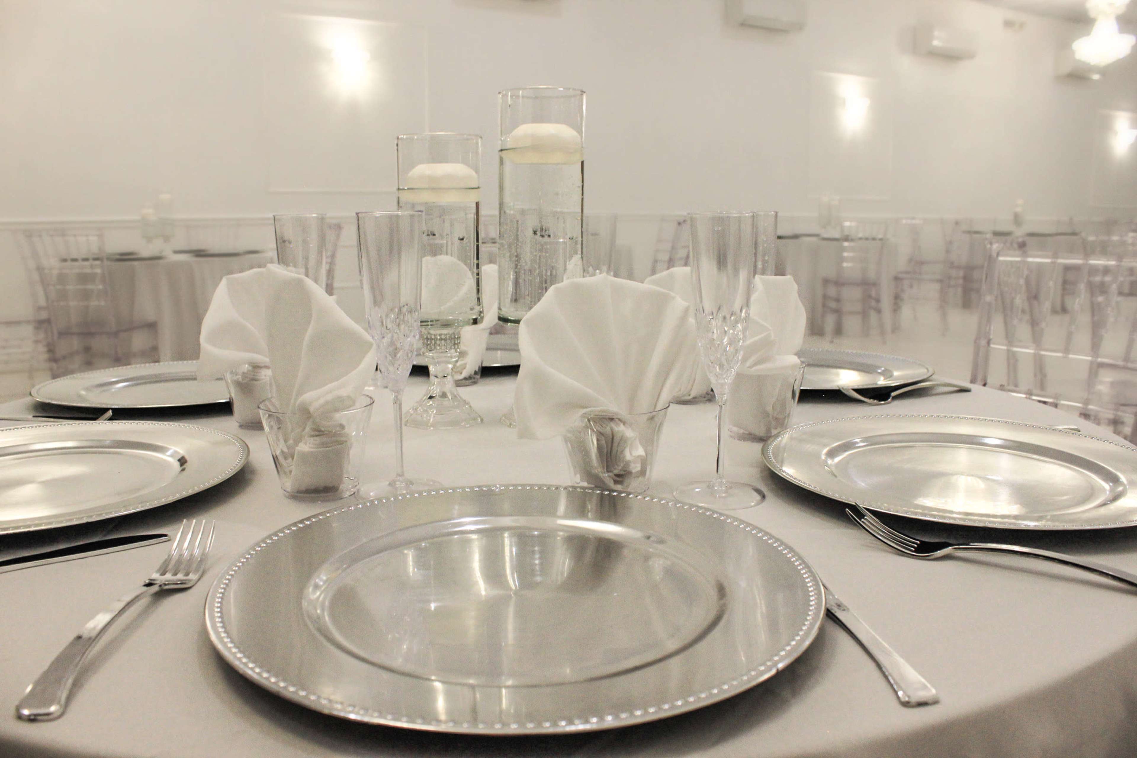 An elegantly arranged dining table set with silver chargers, crystal glassware, and folded napkins, with decorative centerpieces in clear vases.