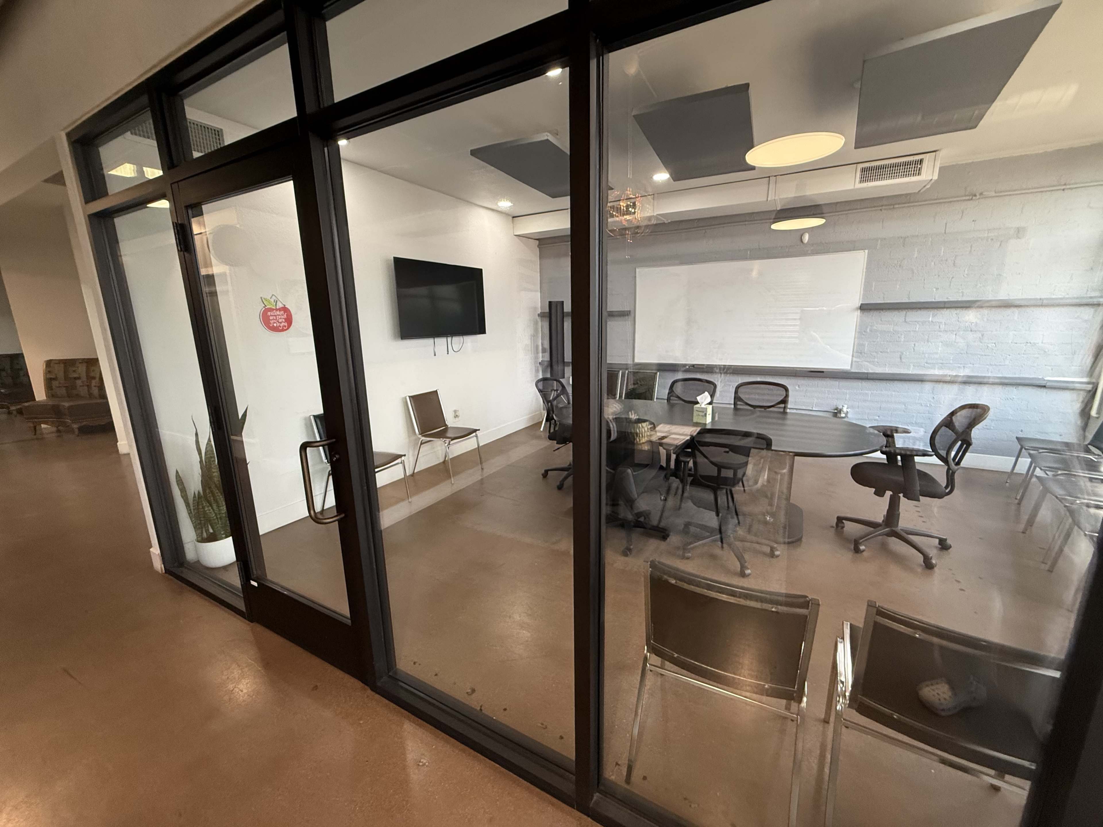 The image shows a glass-walled conference room with several chairs, a table, and a wall-mounted TV.