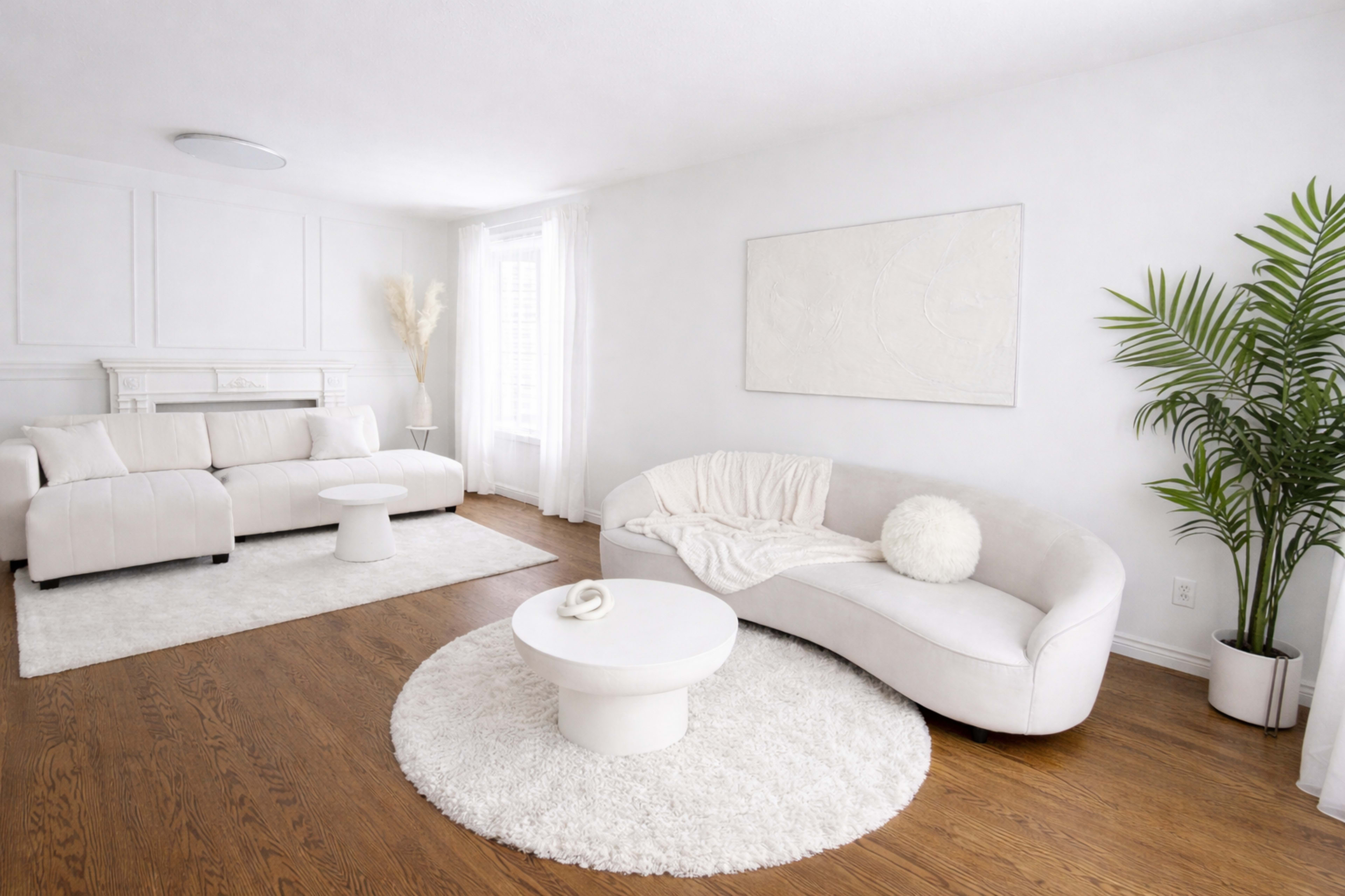 The living room features light-colored furniture, a round coffee table, a large area rug, and a potted plant, all set against white walls.
