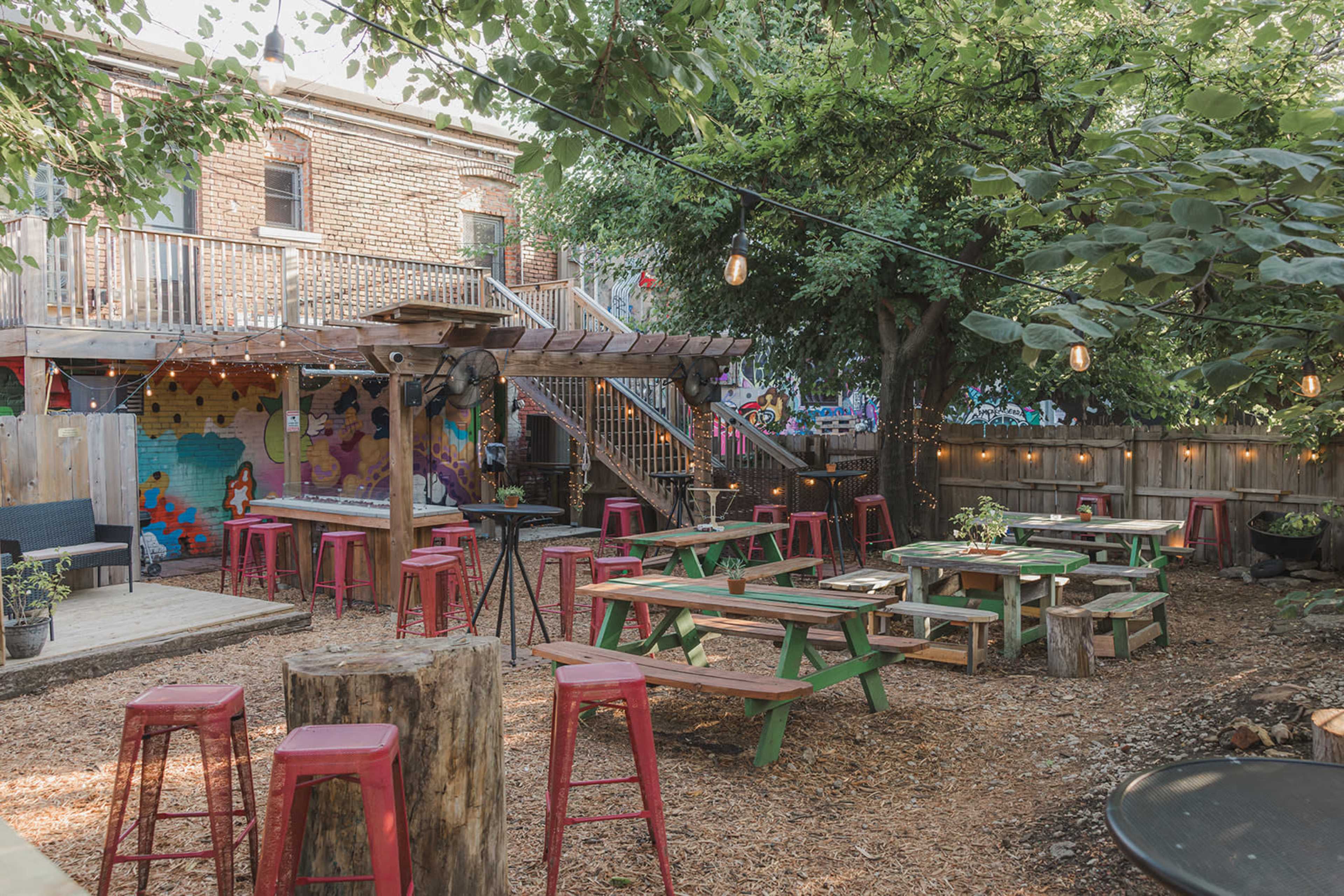 The image shows a cozy outdoor patio area equipped with colorful picnic tables and red stools surrounded by trees and string lights.