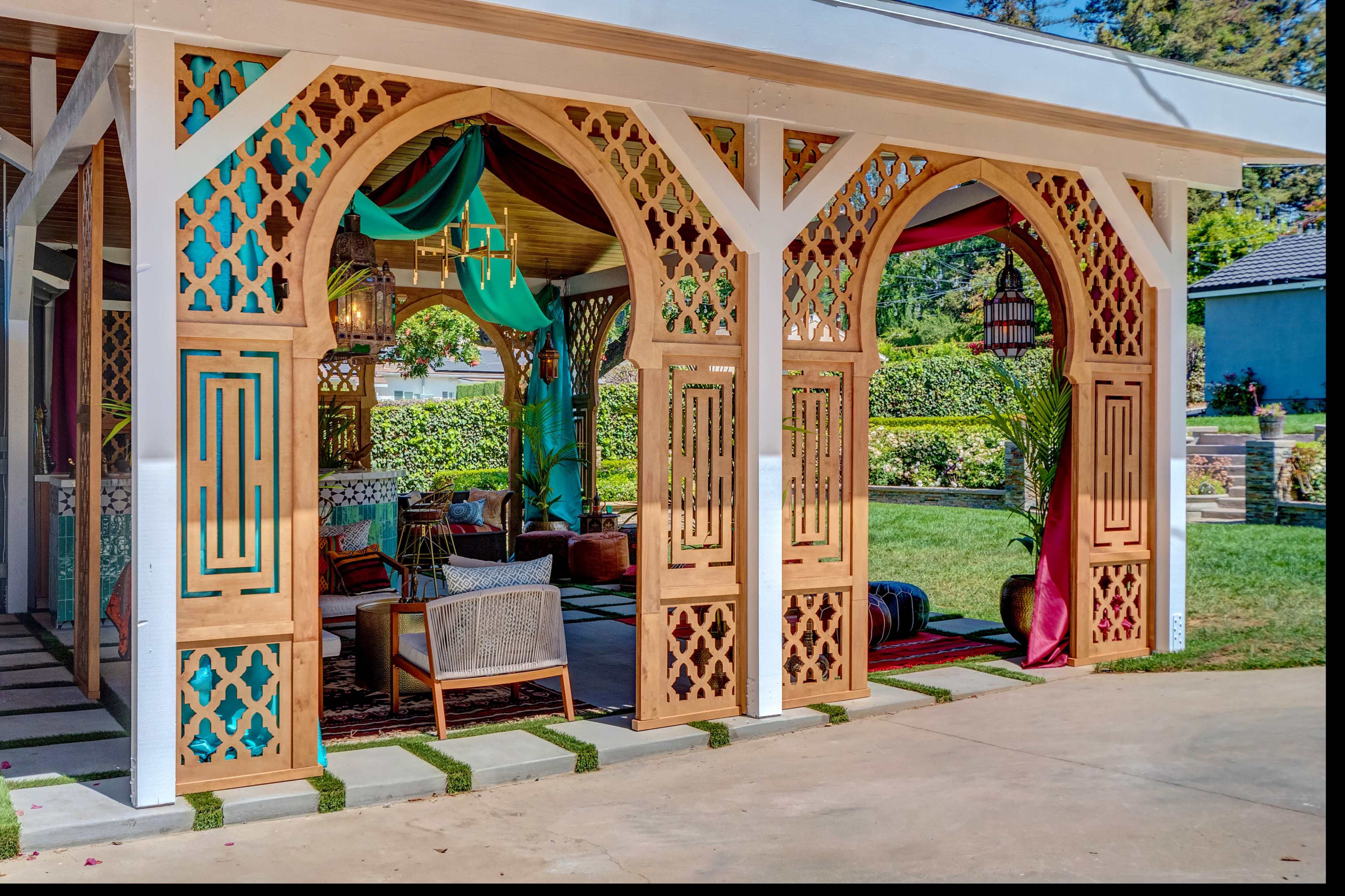 A decorative outdoor pavilion with intricate wooden latticework, vibrant textiles, and comfortable seating arranged in a garden setting.