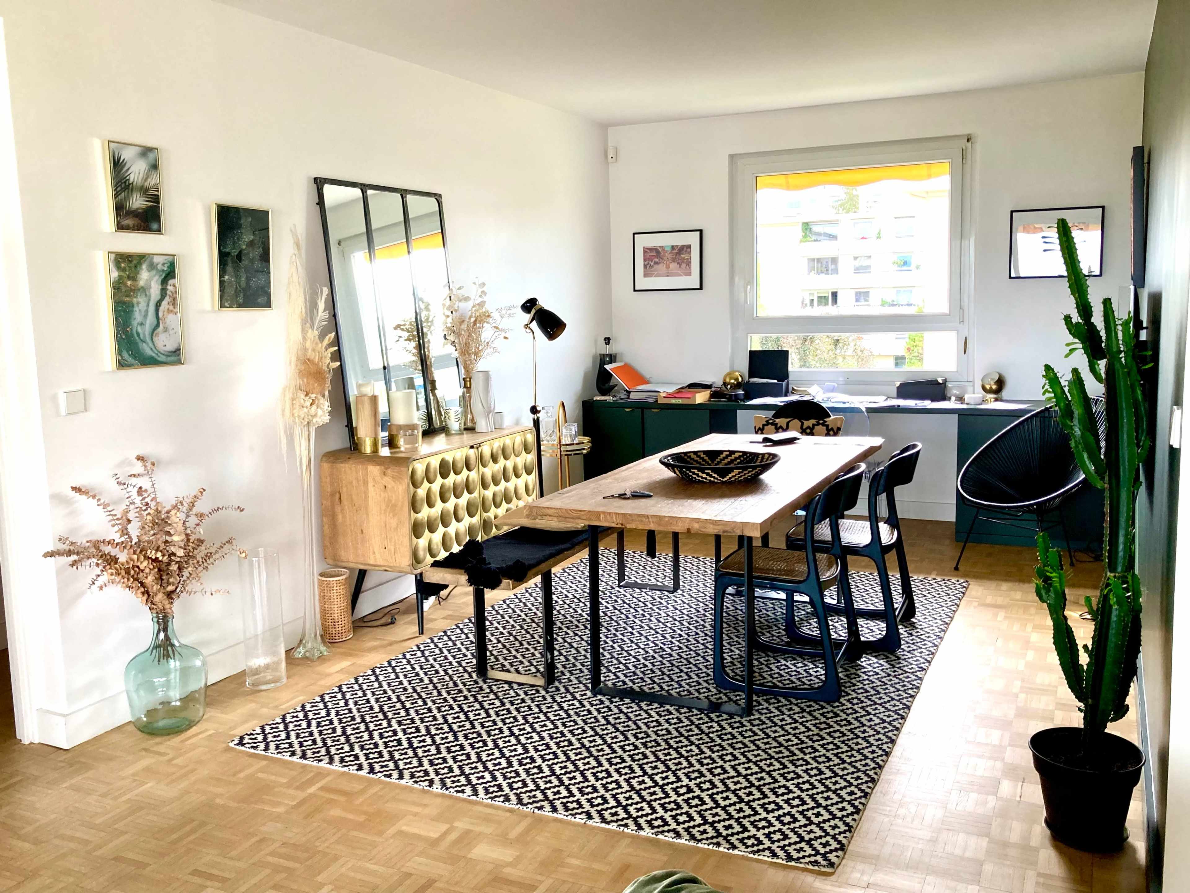 A modern dining area with a wooden table, black chairs, a patterned rug, and decorative plants against a bright, minimalist background.