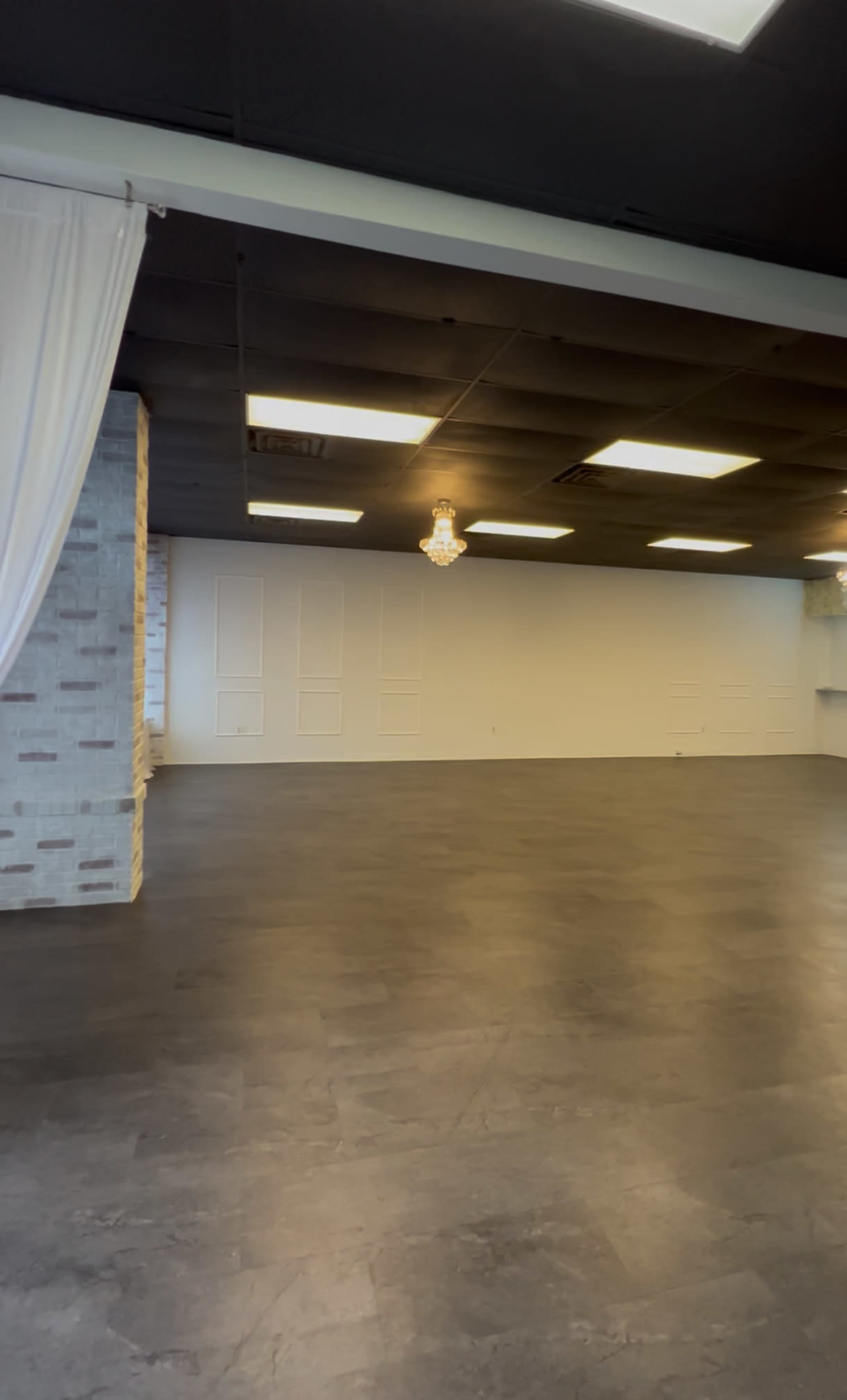 The image shows an empty indoor space with gray flooring, a light-colored brick wall, and a chandelier hanging from the ceiling.