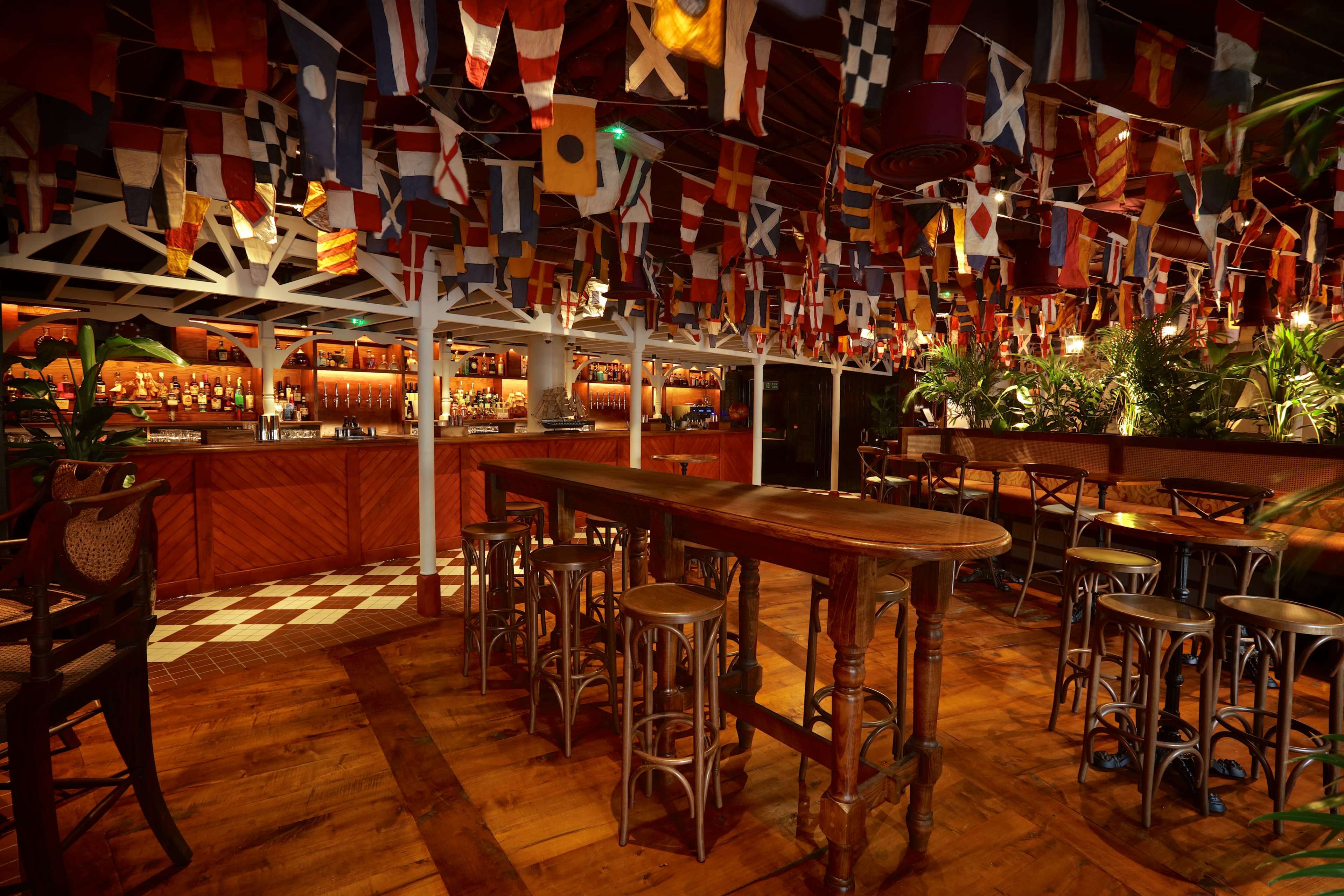 The interior of a bar features a wooden floor, a long table in the center, and colorful flags hanging from the ceiling above a well-stocked bar.