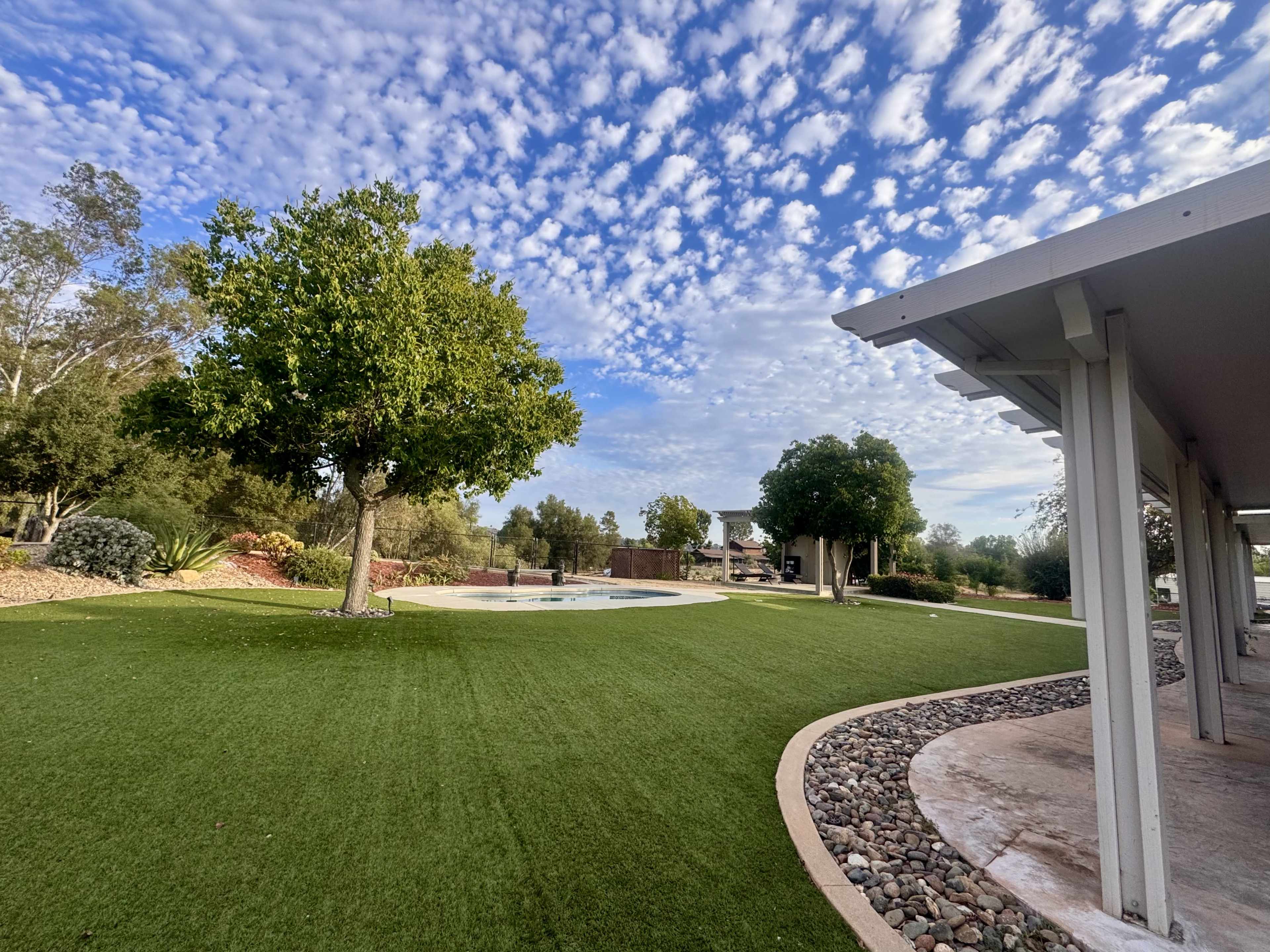 A landscaped yard features a green lawn, a pool, and scattered trees under a cloudy sky.