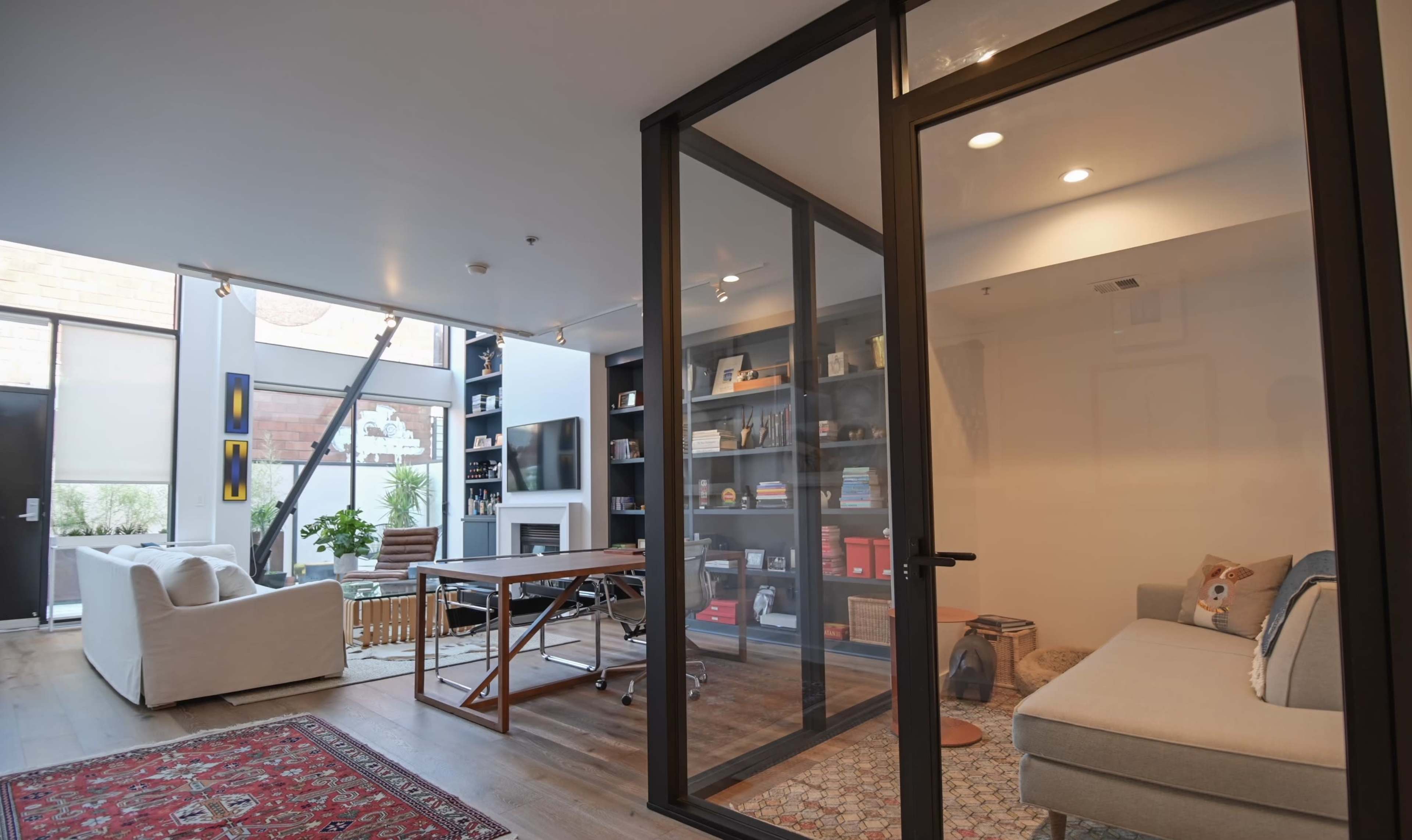 The image shows a modern office space featuring a glass-enclosed room with a desk and bookshelves, adjacent to a lounge area with a couch and a large window.